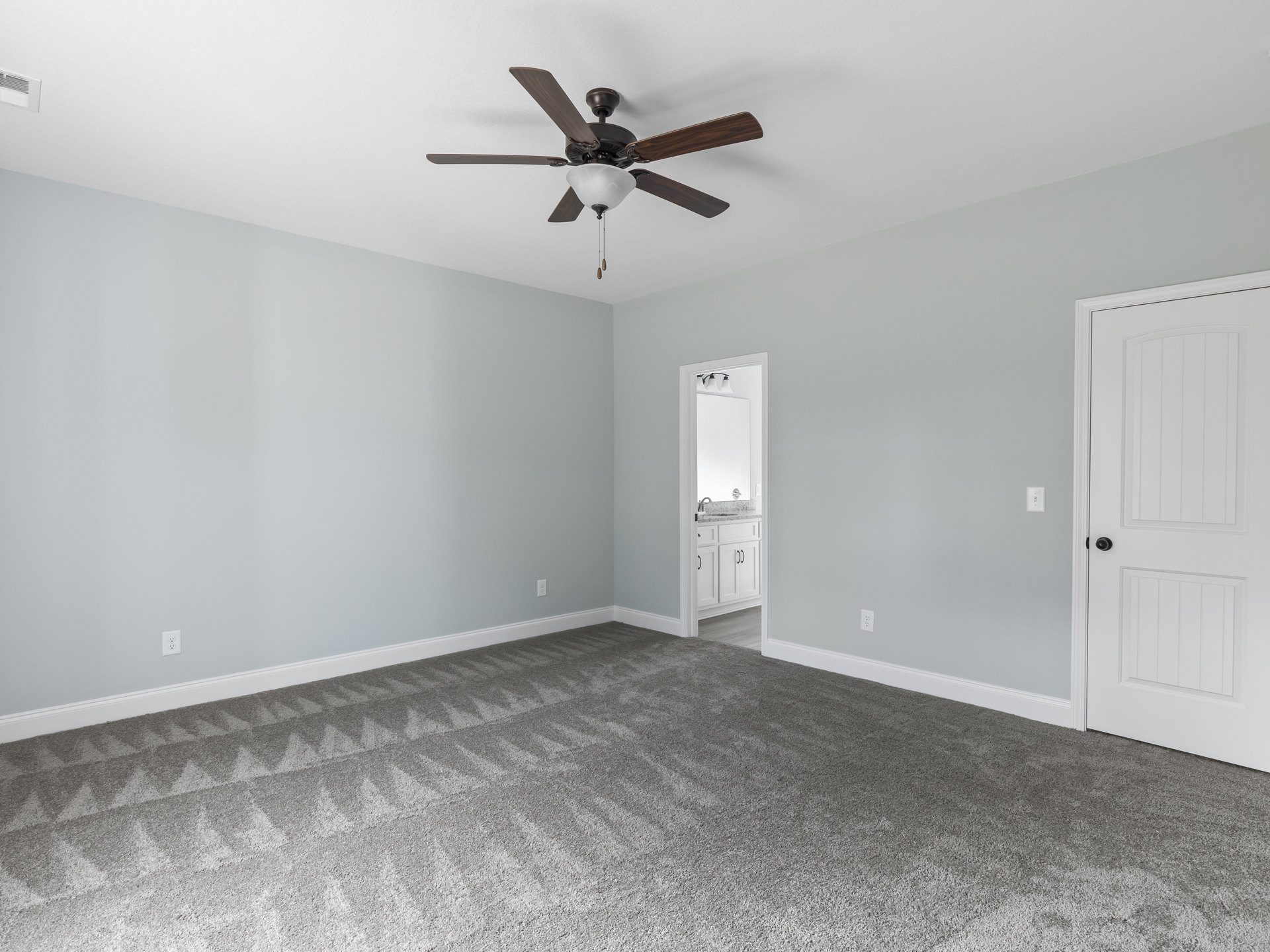 Carpeted bedroom with a ceiling fan and light fixture, white paneled door with black knob, bathroom vanity and mirror visible through open doorway