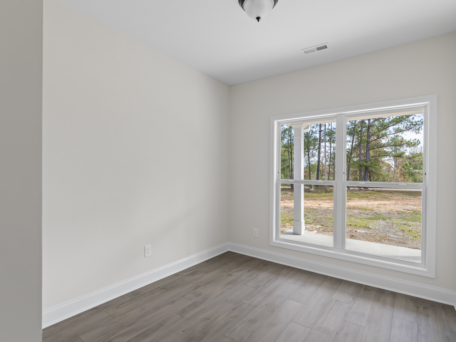 Sunlit room with white walls and ceiling, large window overlooking trees and dirt field, wood laminate flooring with white trim, recessed ceiling light