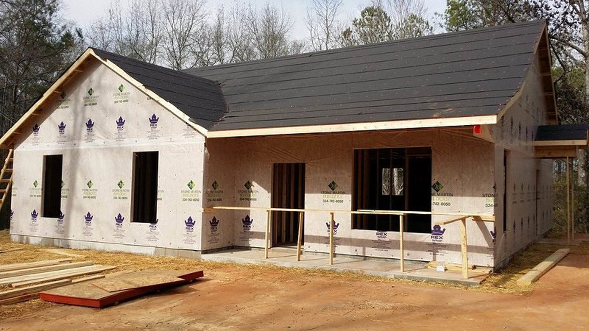 Wood-framed house under construction with exposed beams, partially enclosed porch, and unfinished siding, surrounded by trees and a temporary fence