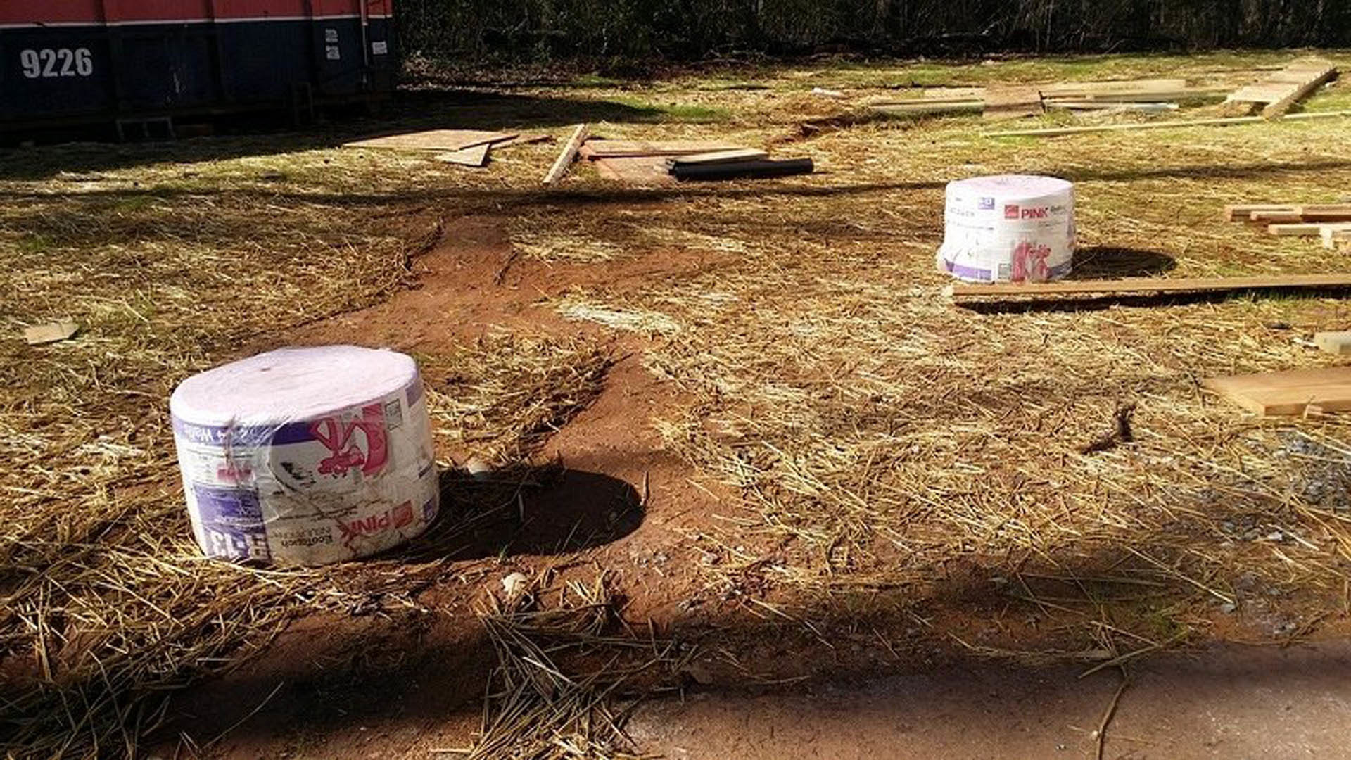 Several rolls of paper scattered on grassy ground near a red and blue wall, with trees and waste containers in the background.