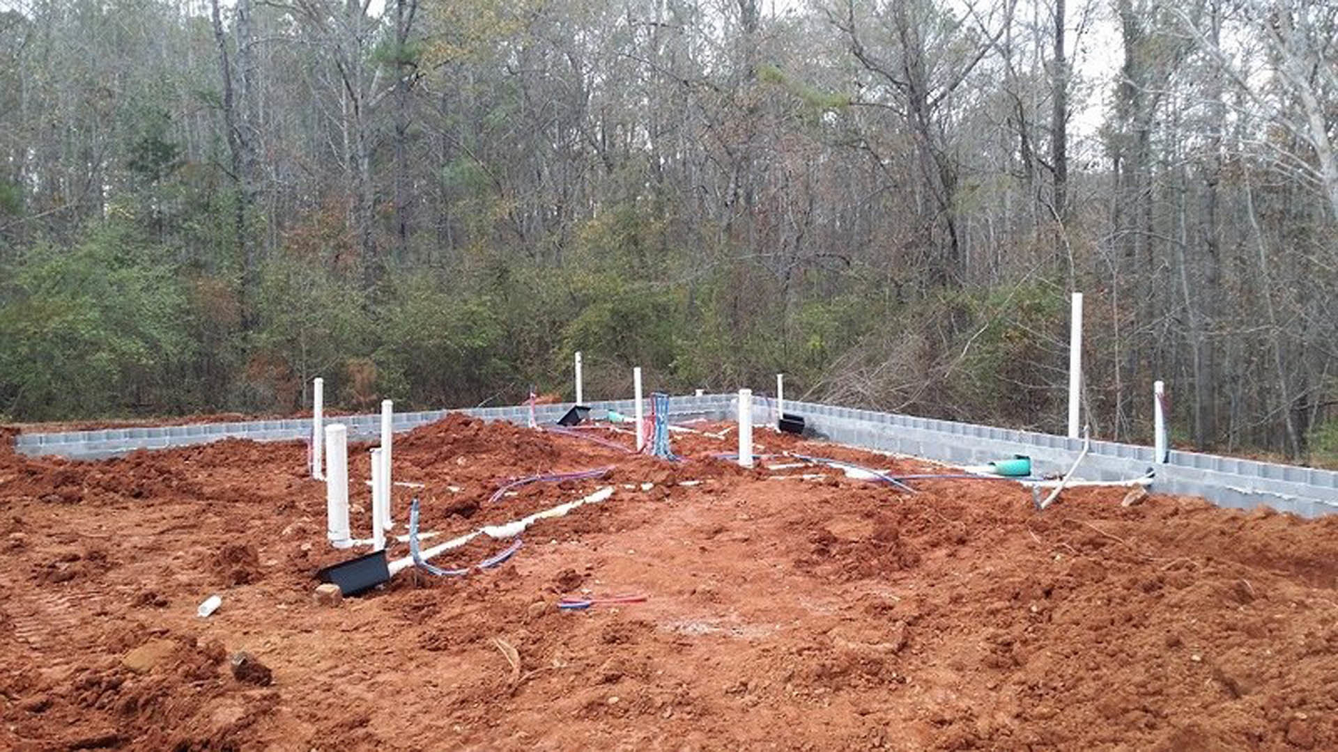 Red dirt construction site with white foundation poles, surrounded by trees and soil.