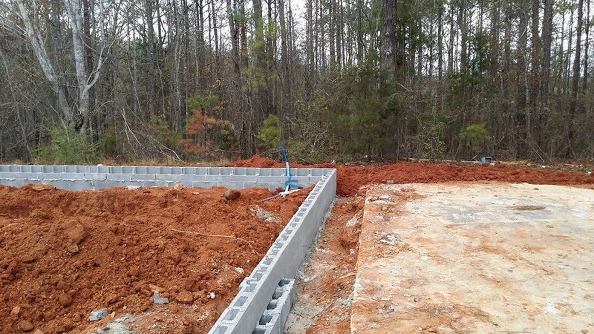 Concrete foundation surrounded by red dirt, bare trees in the background, outdoor construction site.