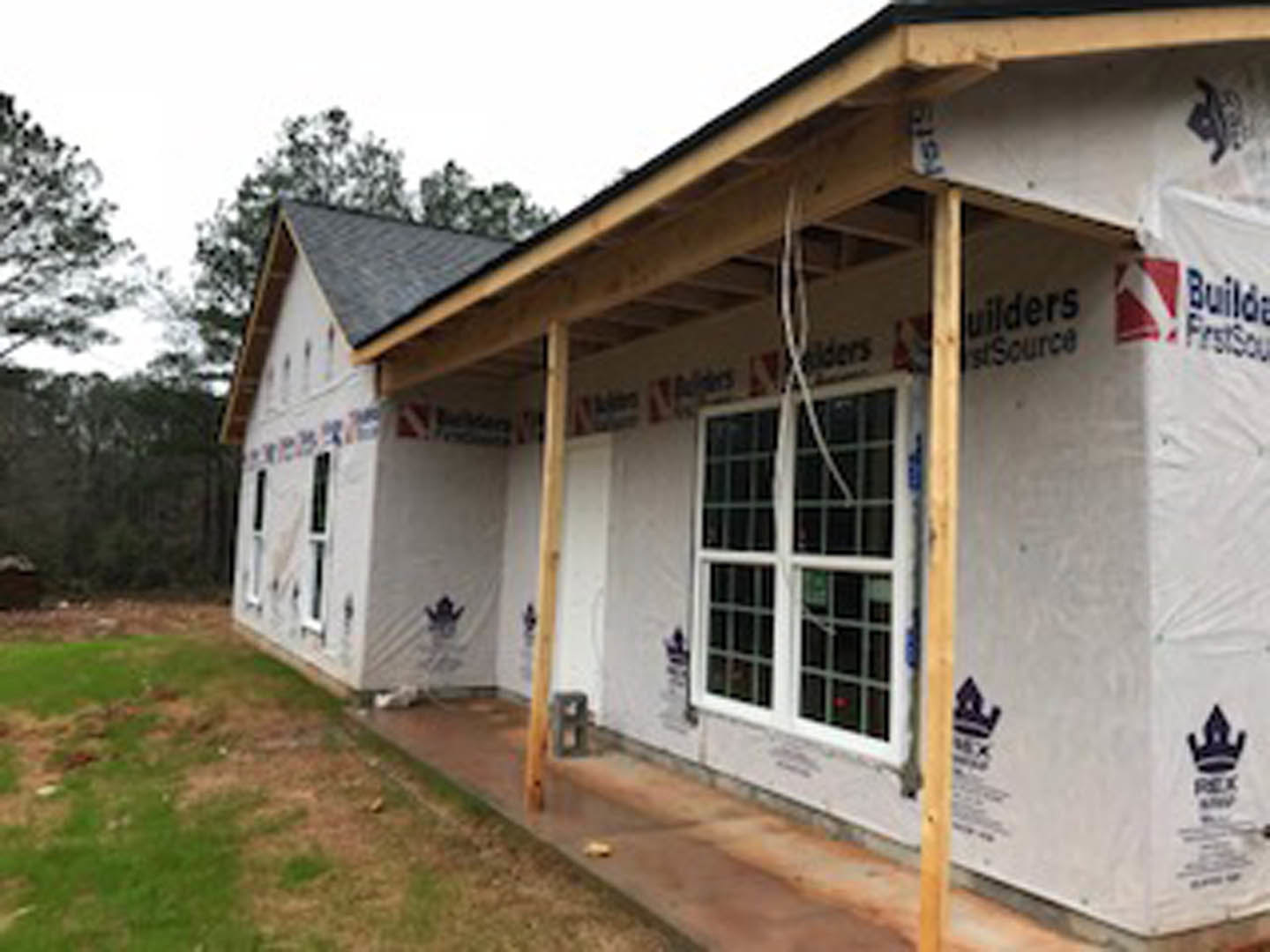 Wood-framed house under construction with covered porch, exposed beams, white siding, and black trim; grassy yard and trees in background