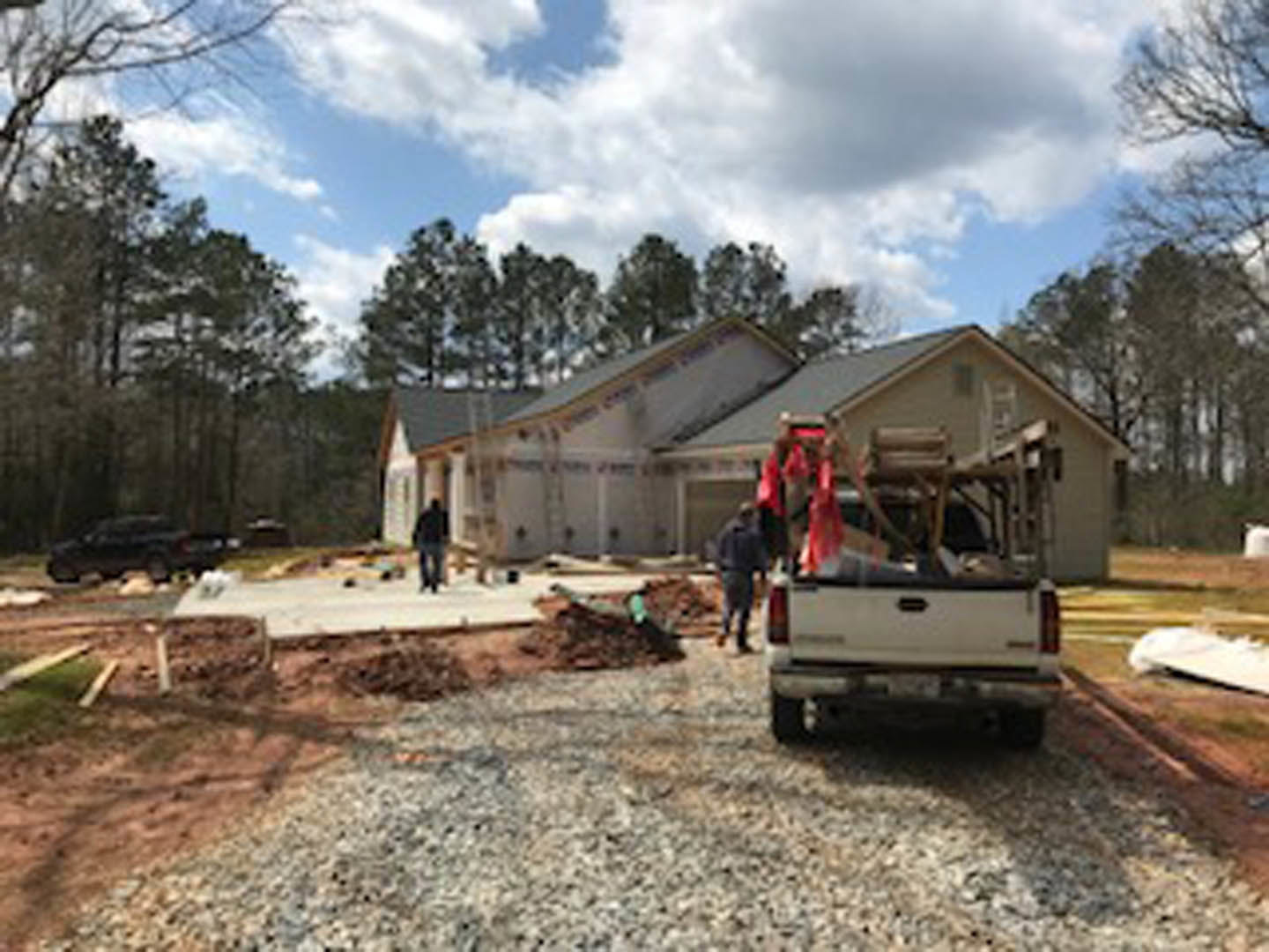 Partially built house framed with exposed wood, white truck parked in front, two people standing near construction materials, orange traffic cone on gravel driveway, trees and