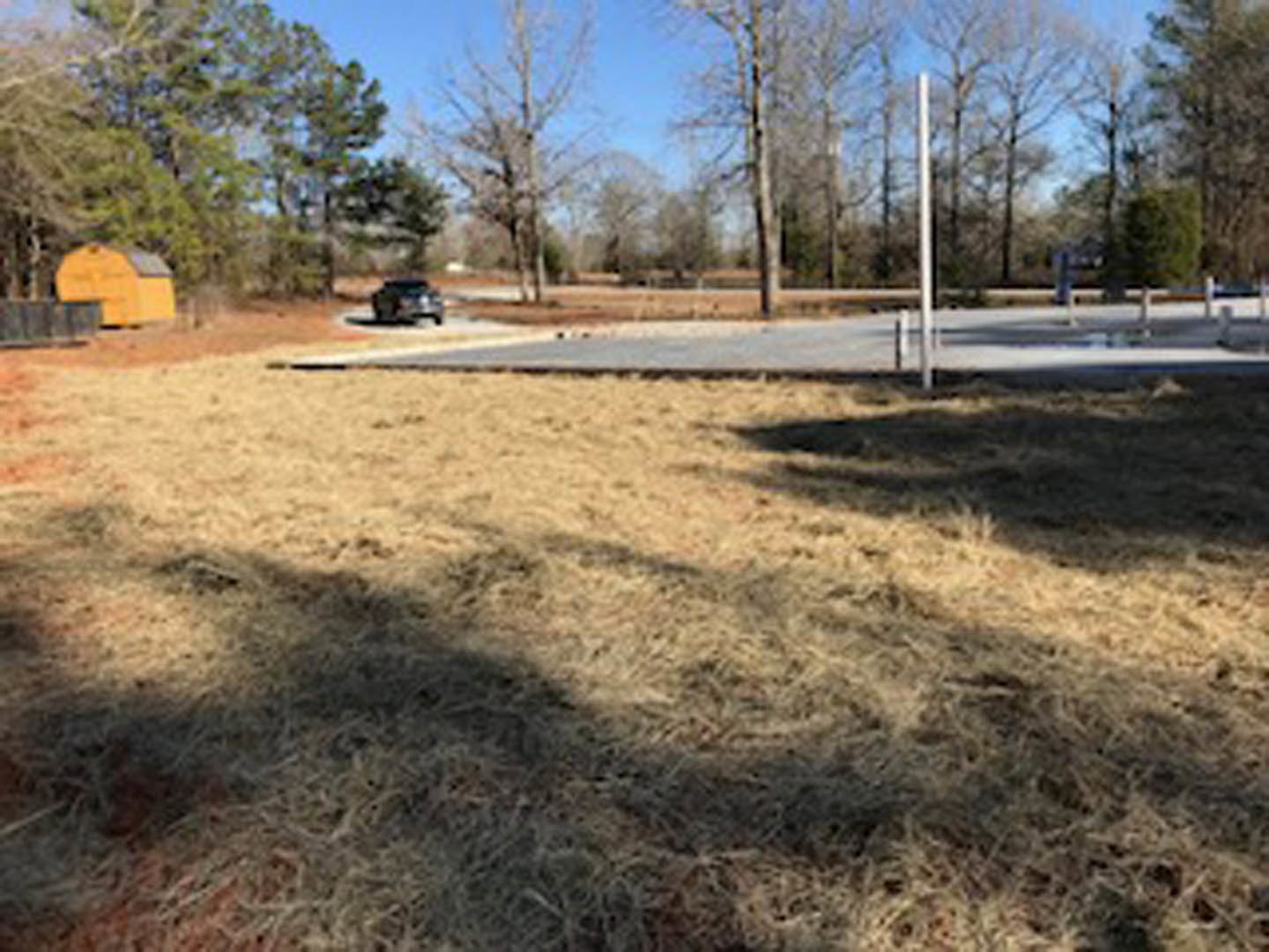 Parking lot bordered by grass and mature trees, with a yellow barn featuring a blue door in the background; sunlight casts shadows across the grassy field.