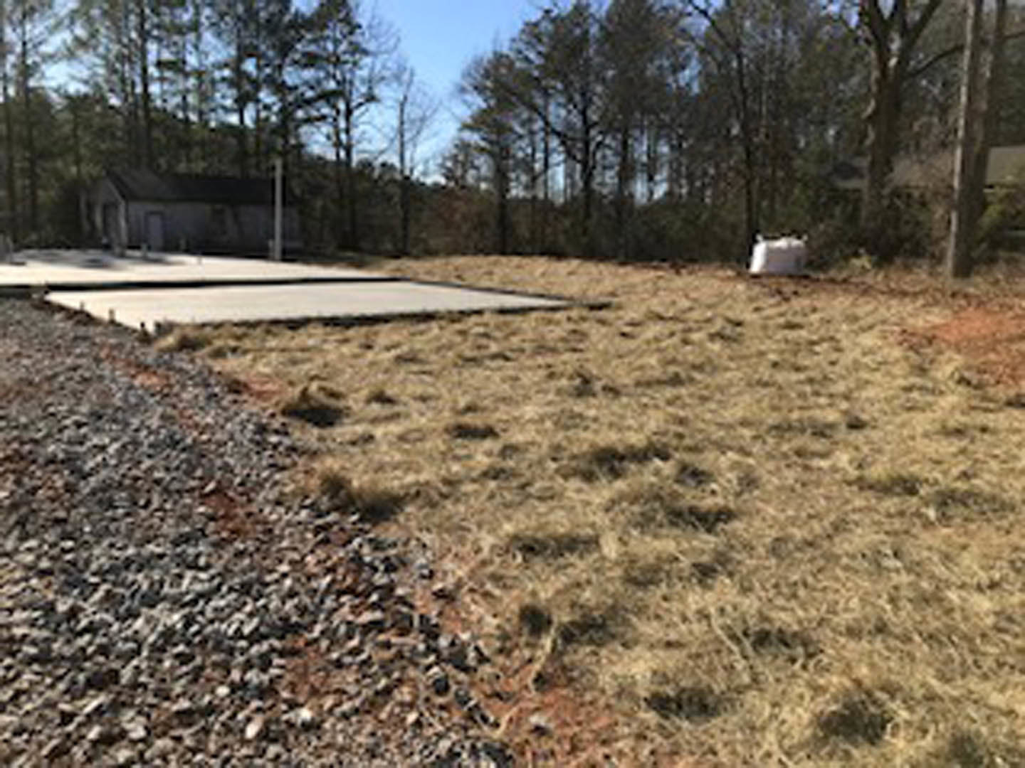 Modern house with concrete slab foundation, gravel driveway, grassy yard, and large windows; trees and open sky in background.