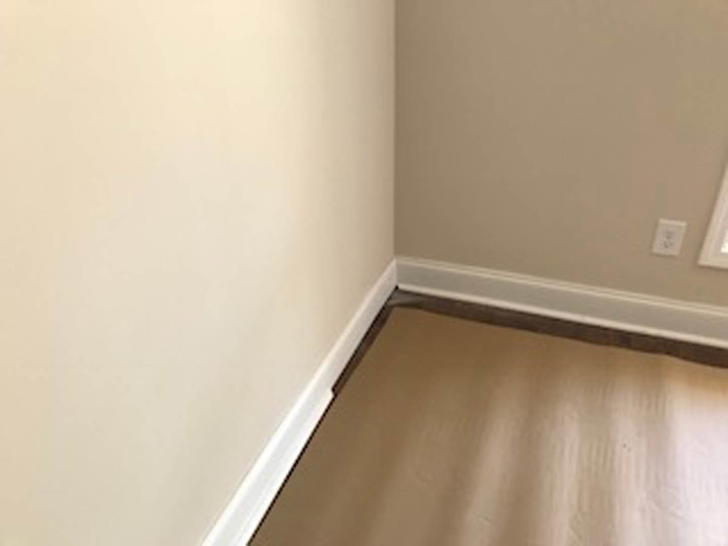 Corner of a room featuring brown hardwood flooring, white baseboard, and smooth white plaster wall with a visible electrical outlet