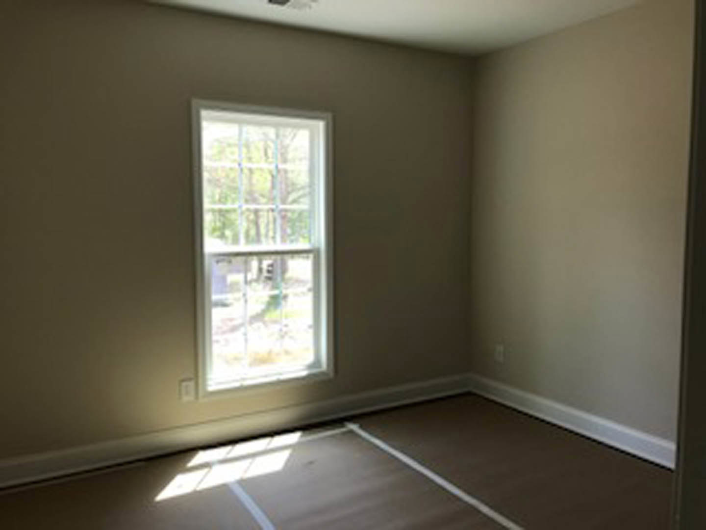 Sunlit room featuring a large window with a view of trees, smooth wood flooring, white plaster walls, and a visible light switch near the doorway