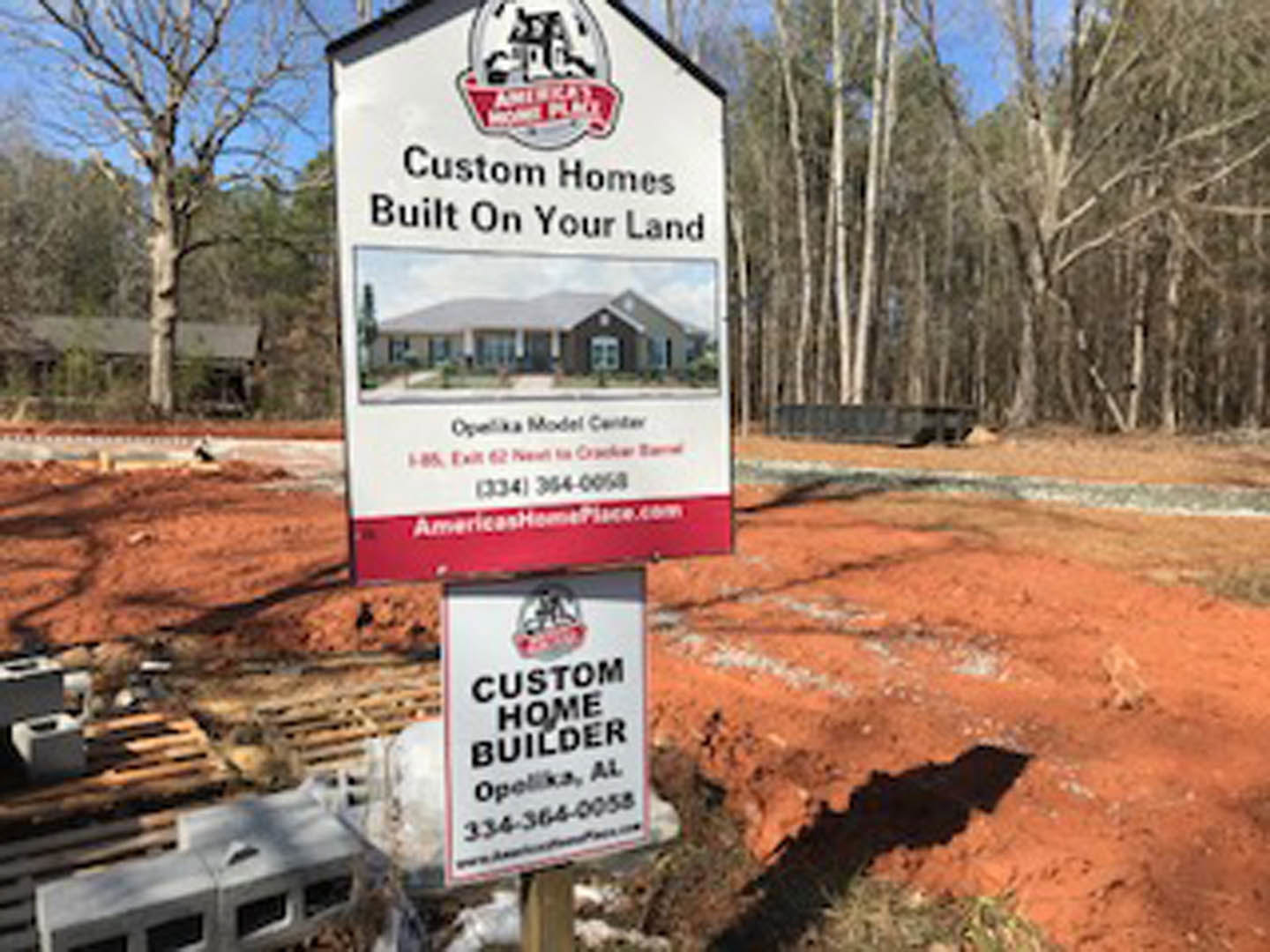 Wooden post-mounted sign with house graphic stands on dirt ground, surrounded by trees and open sky