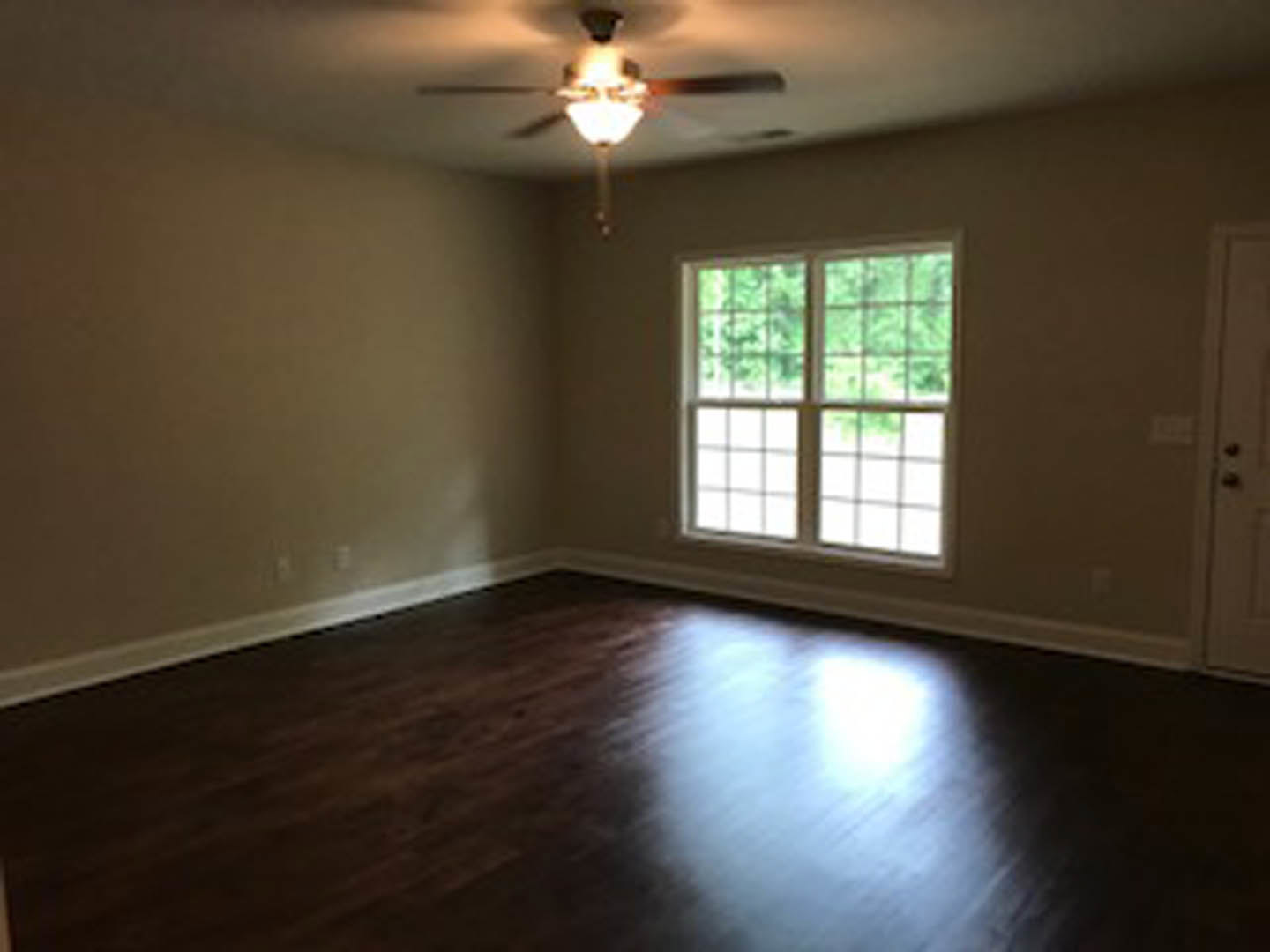 Dark wood flooring with white walls, ceiling fan, and large window featuring multiple square panes, natural light reflecting across the floor.