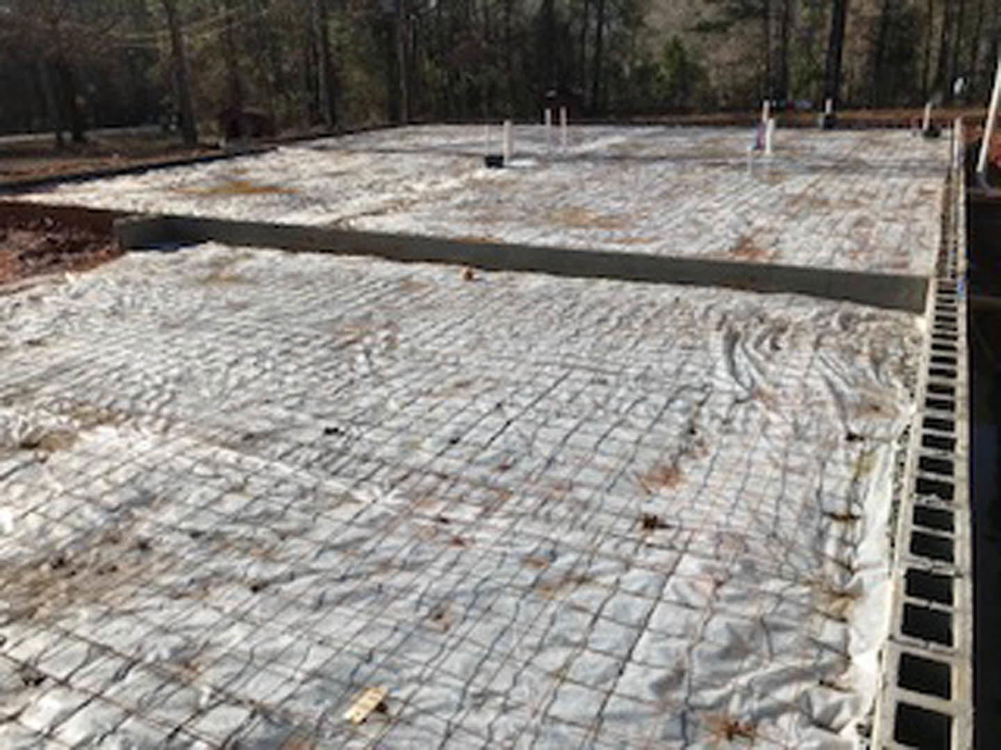 Concrete foundation under construction, covered with a blue tarp, surrounded by dirt and trees, with a temporary fence in the background