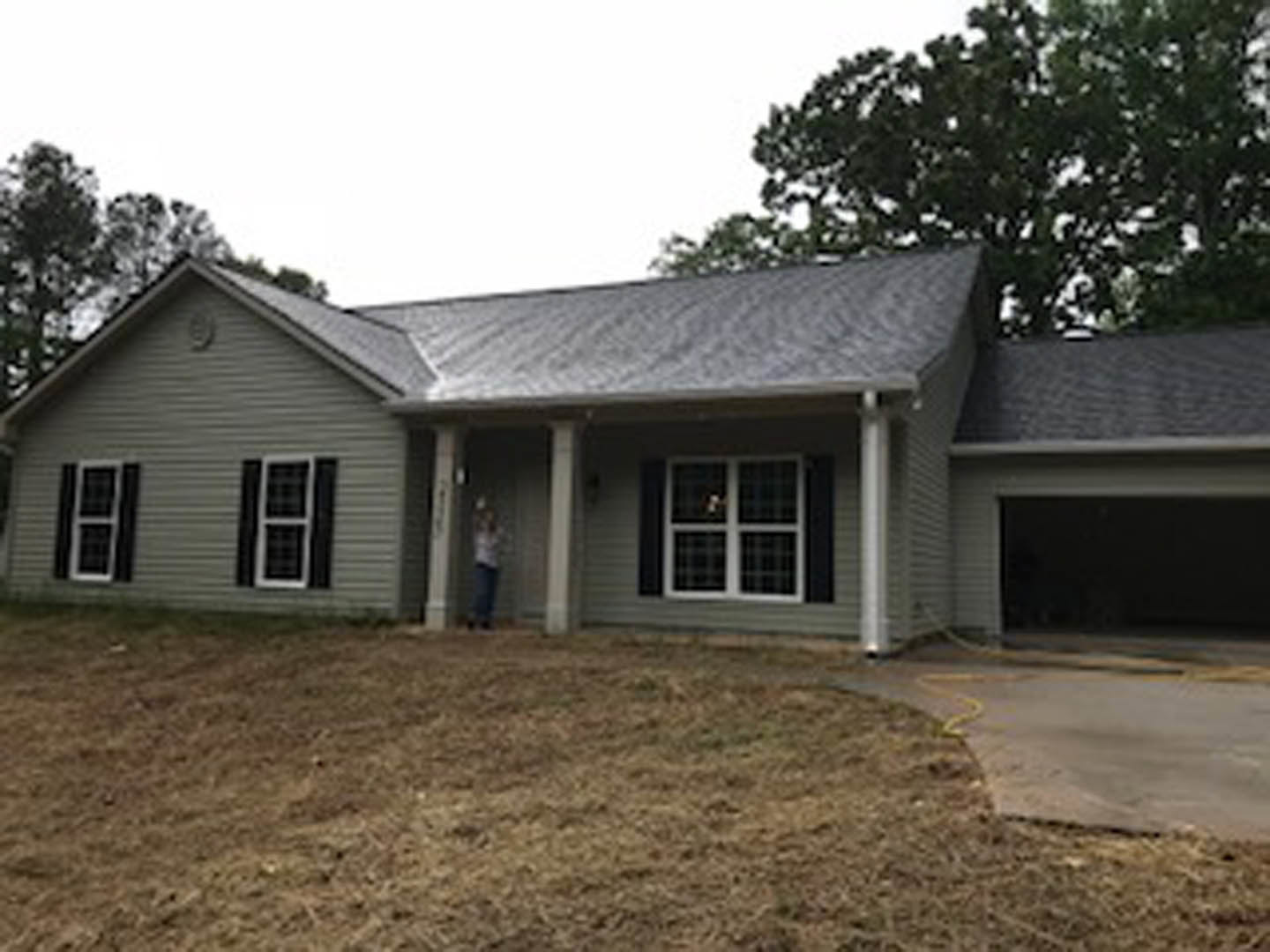 Two-story home with white-framed windows, gray siding, and shingle roof, concrete driveway bordered by green lawn, mature tree on the left, person standing near front entrance