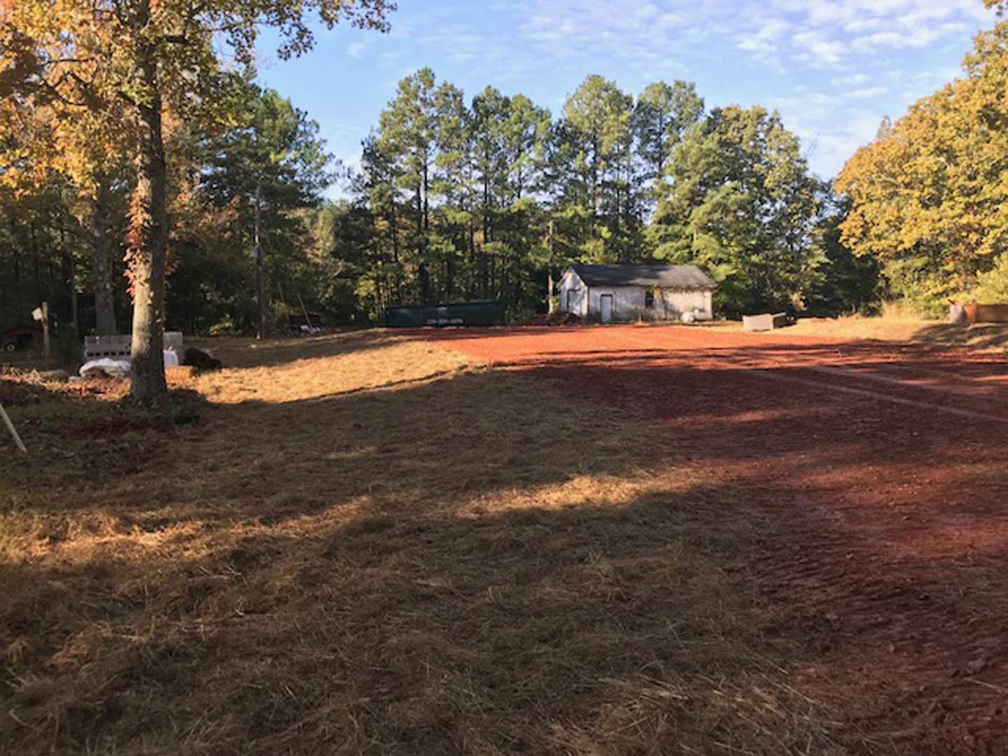 Dirt road bordered by grass field, mature trees with yellow leaves, and a distant custom home partially visible against a blue sky