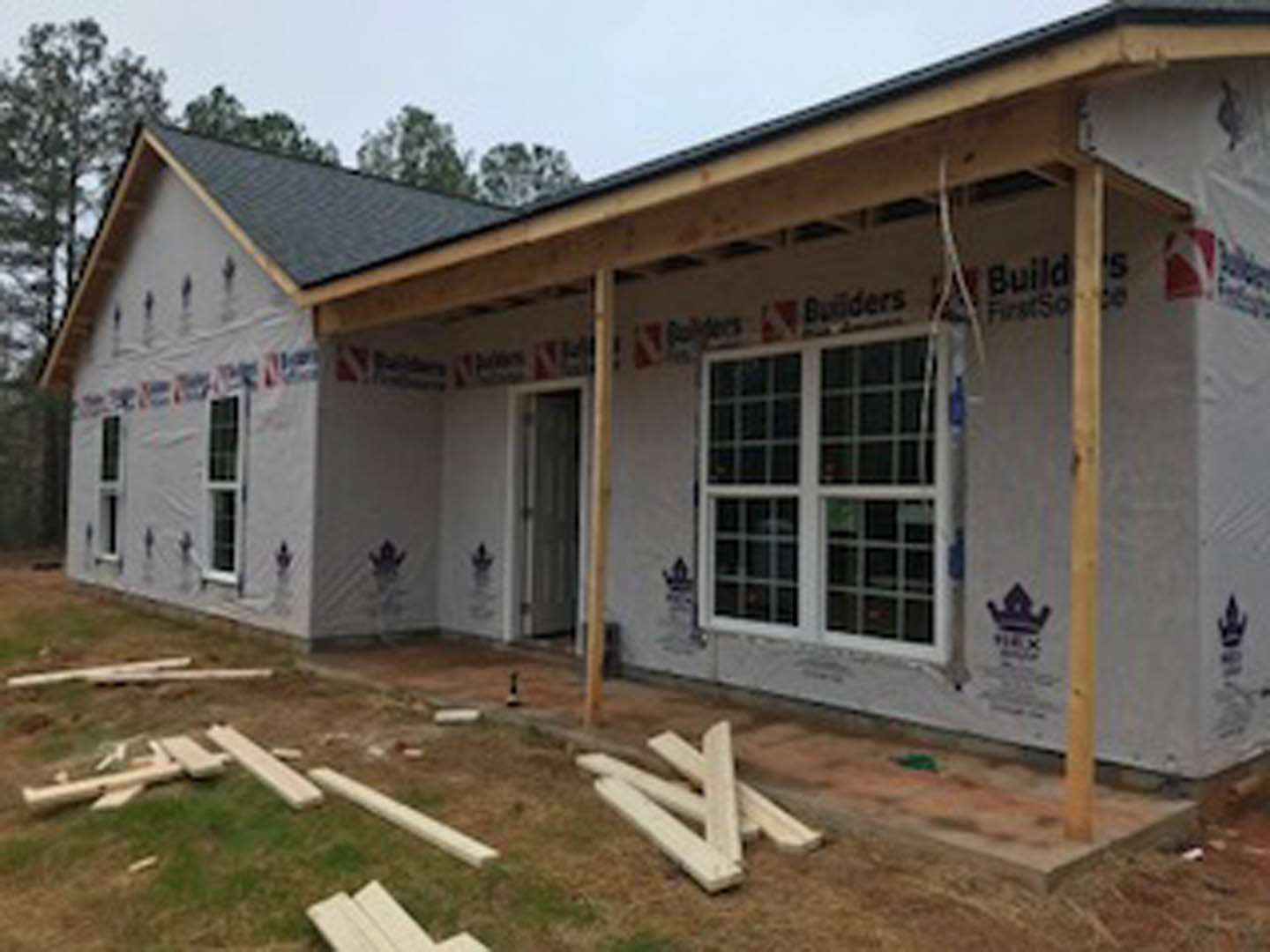 Wood-framed house under construction with exposed siding, stacked lumber on the ground, and partially installed windows and doors