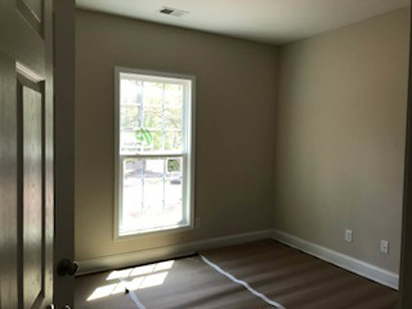 Carpeted room with a wooden floor border, white plaster walls, large window showing greenery outside, white electrical outlet, and a door on one side