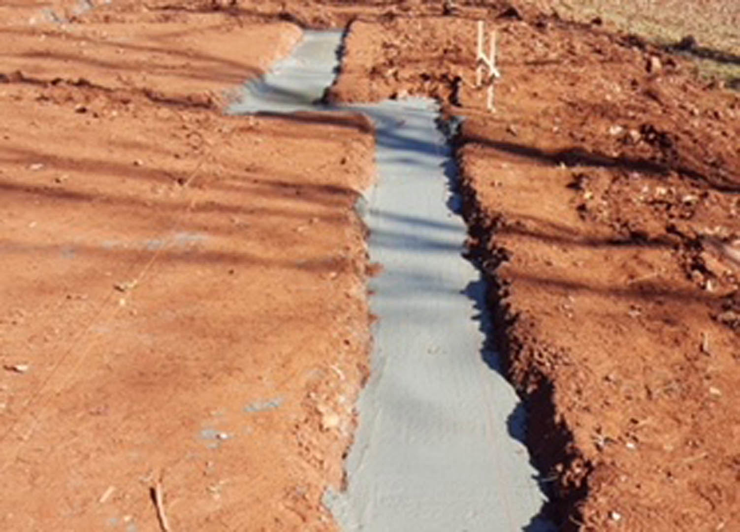 Wide view of a deep trench dug into brown soil on a residential lot, surrounded by loose dirt and sparse vegetation