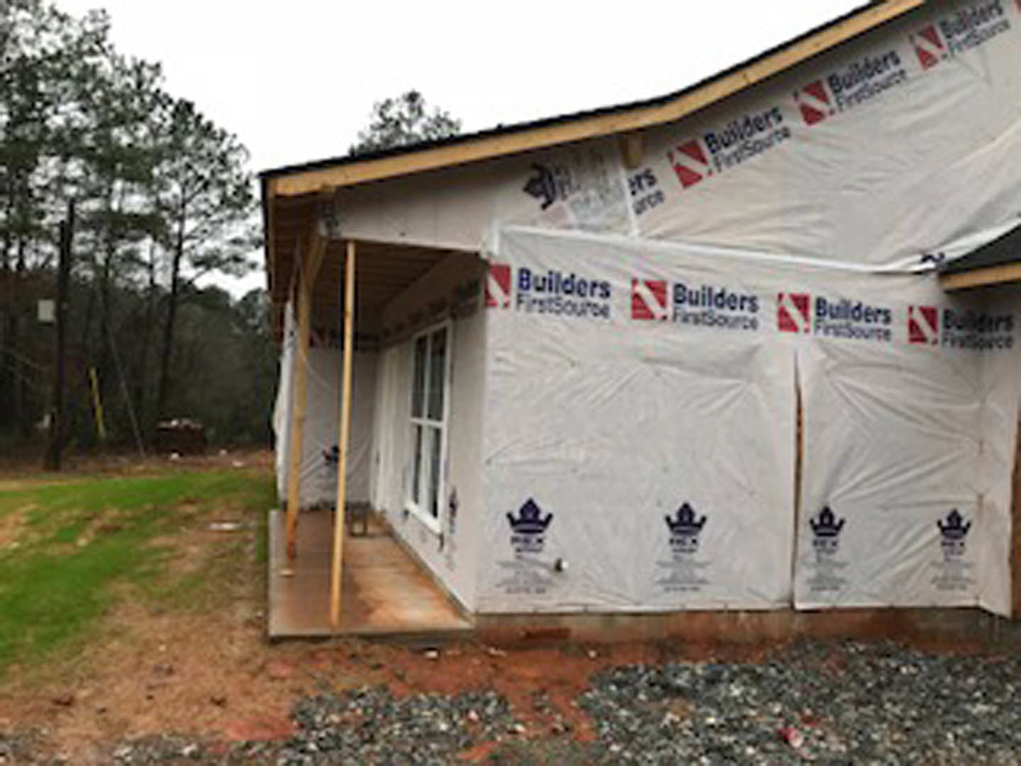 Two-story house under construction with white plastic sheeting covering exterior walls, unfinished roof, and surrounding trees in background