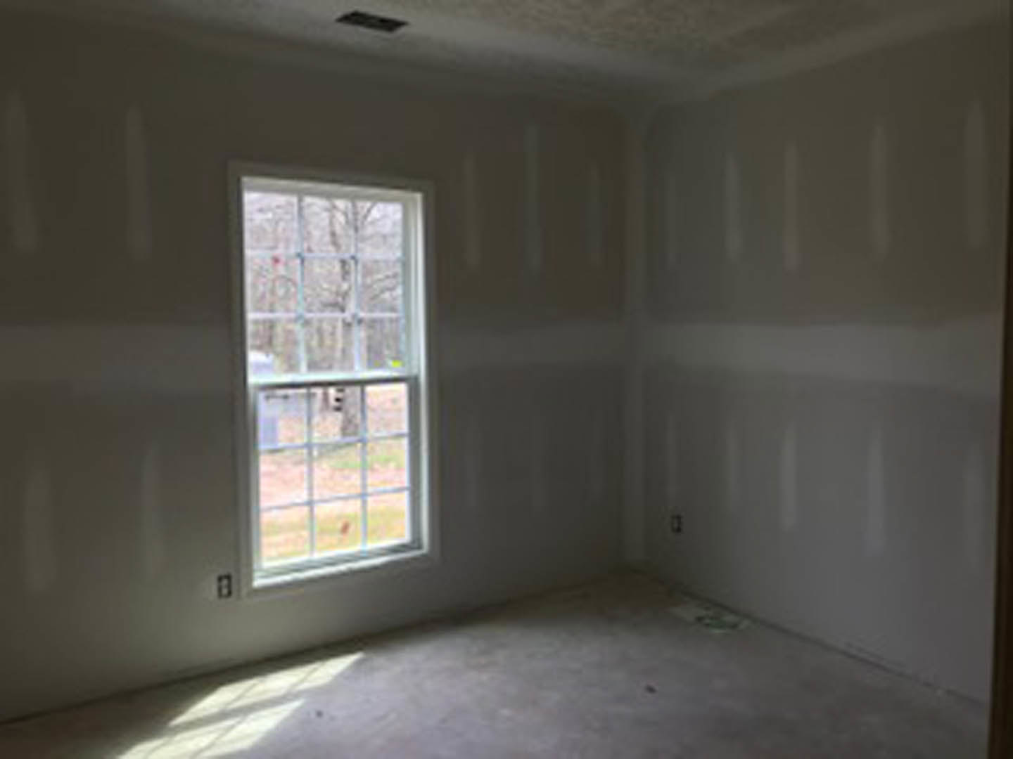 White-walled room featuring two large windows with views of leafy trees, light-colored flooring, and a dark rectangular fixture.