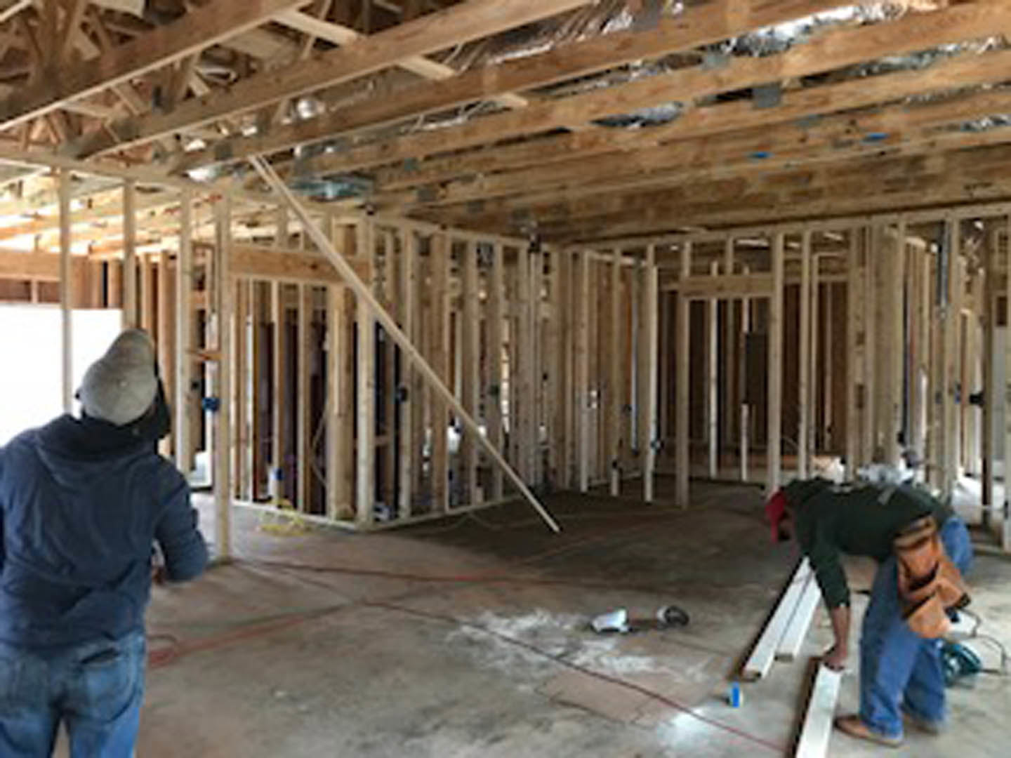 Men inspecting unfinished room with exposed wooden beams, insulation, and construction materials; one wears a blue jacket, another stands on subfloor, faces partially blurred.