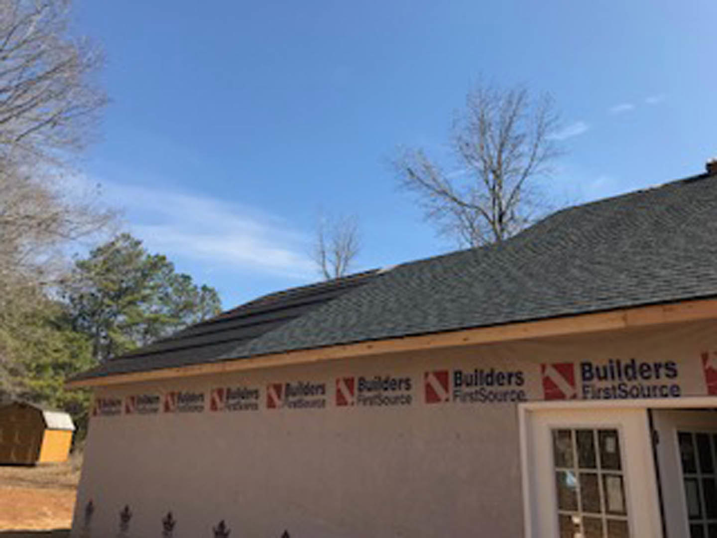 White custom home with gray roof, large front window, and a sign mounted on the side wall; blue sky overhead, tree partially visible at edge of frame.