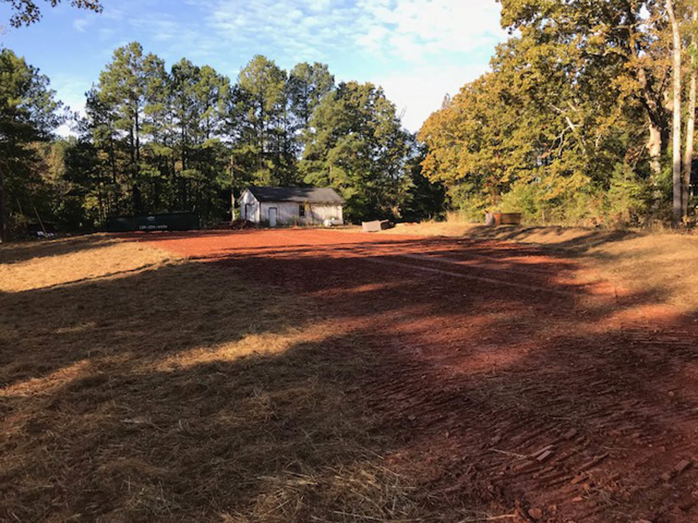 Dirt field with tire tracks bordered by trees, white house with gabled roof in the background under cloudy sky