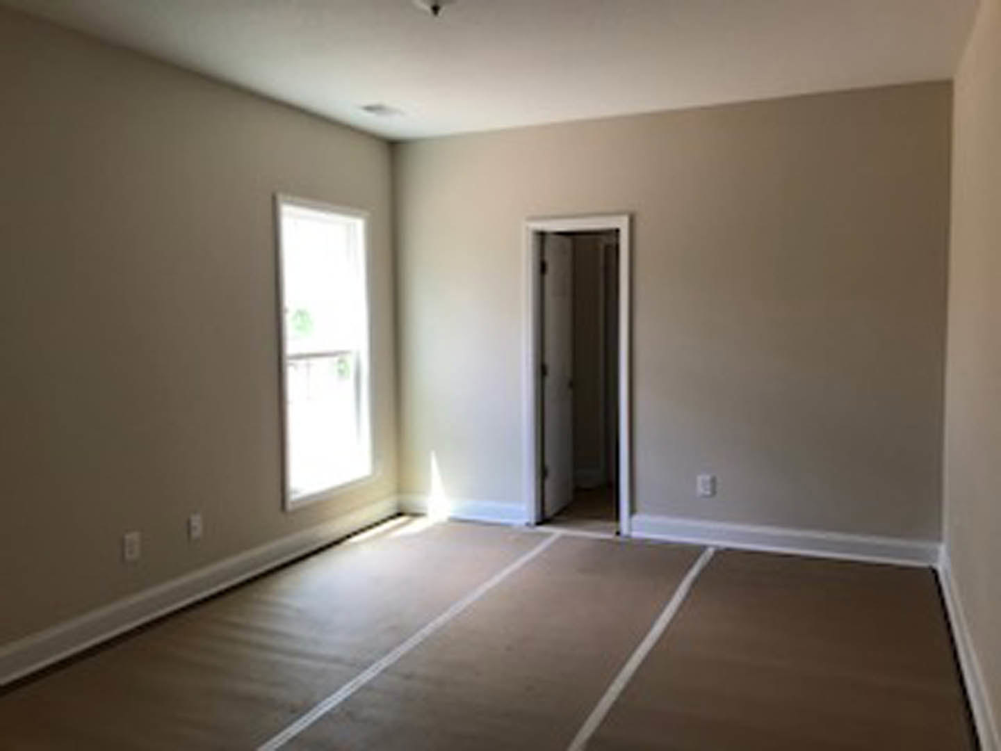 Open white door and large window with white frame in a room featuring light wood laminate flooring and smooth plaster walls.