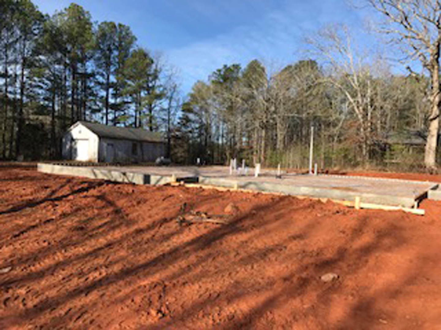 Two-story house with light siding and dark roof, surrounded by mature trees, dirt driveway, and concrete walkway leading to entrance
