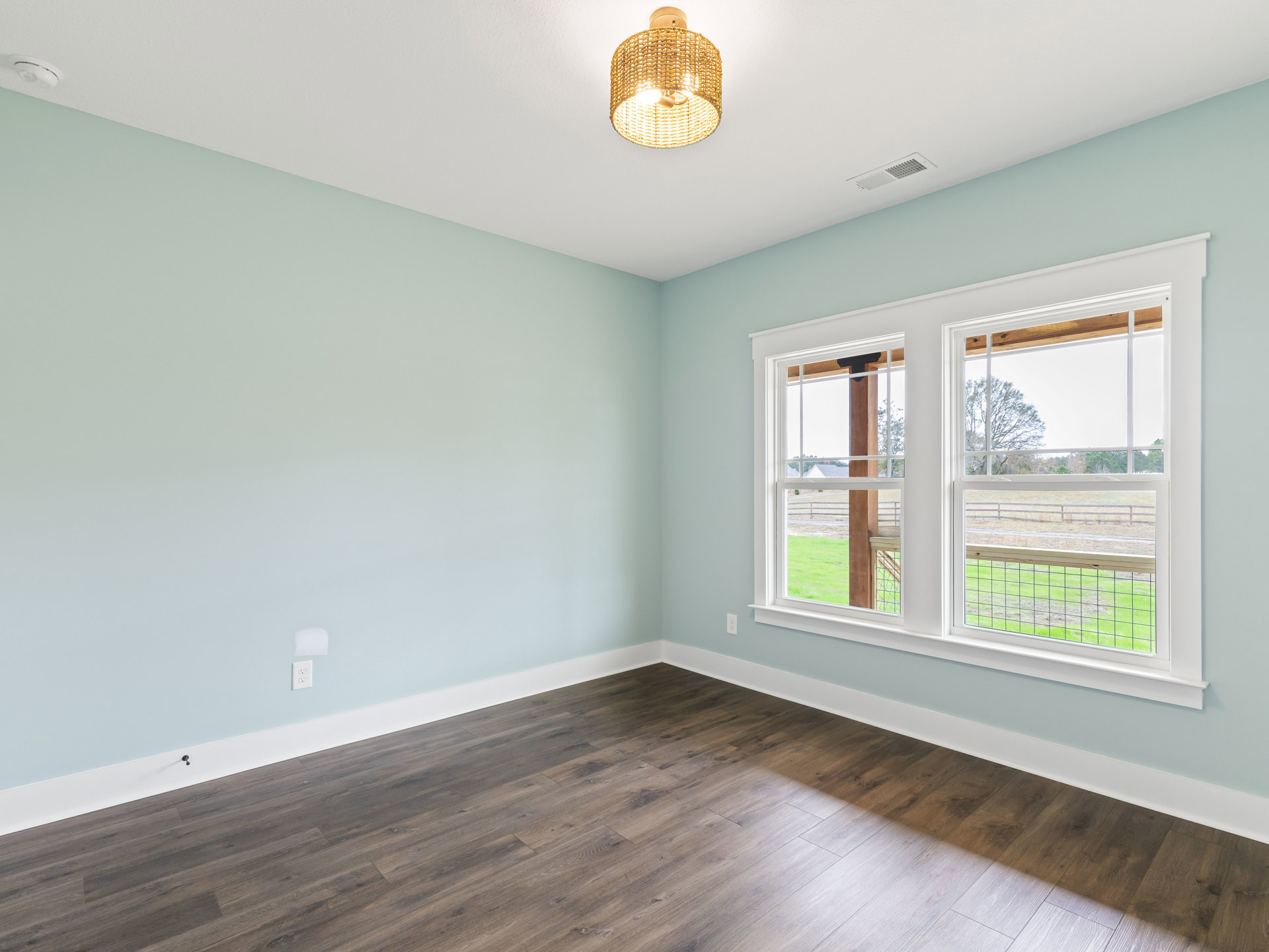 Woven pendant light hangs above wood flooring with white trim, large windows reveal fenced yard and green grass outside