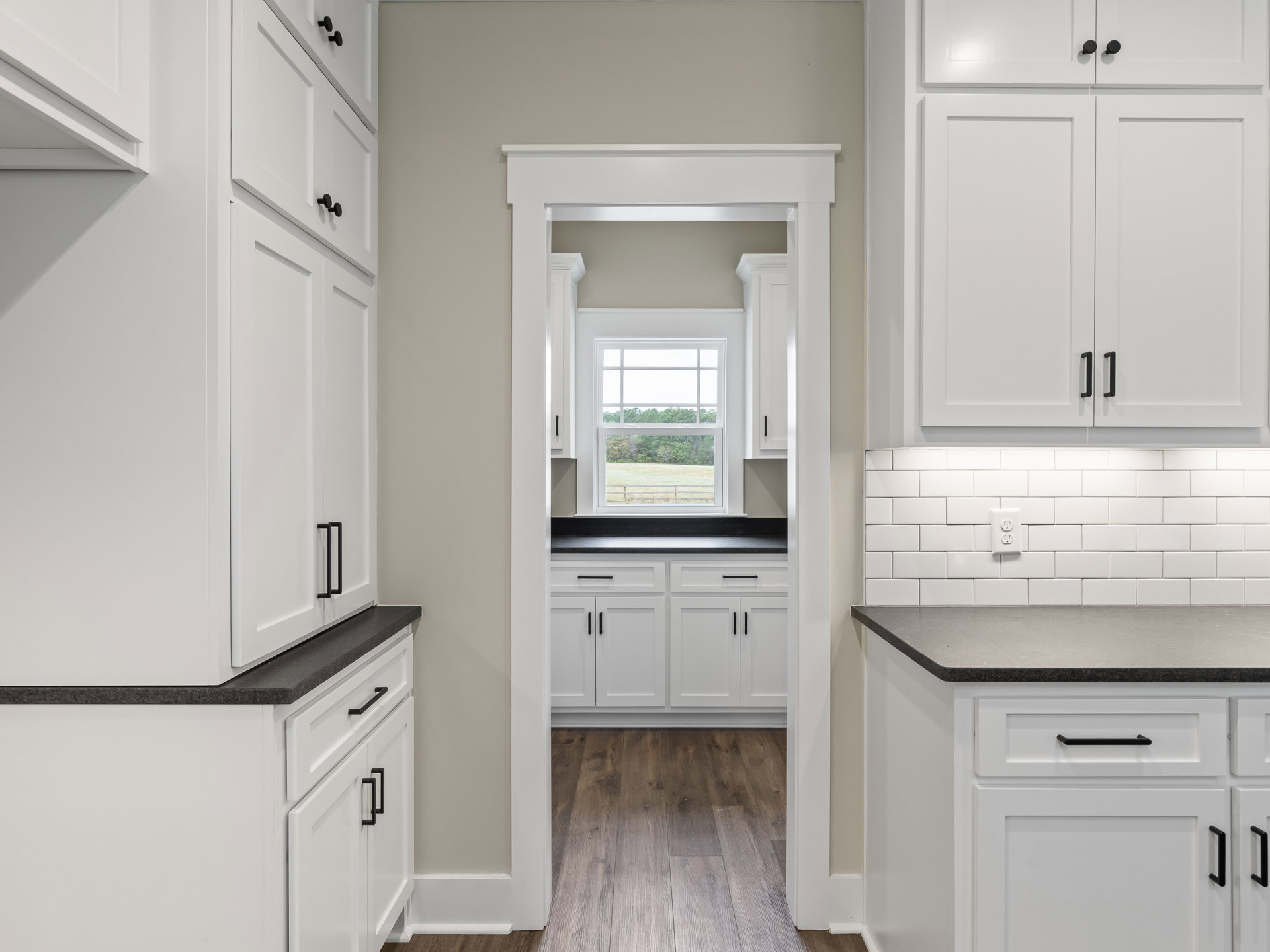 White kitchen cabinets with black countertops, stainless steel sink, wood flooring, window overlooking trees, white door with black handle, black cabinet hardware