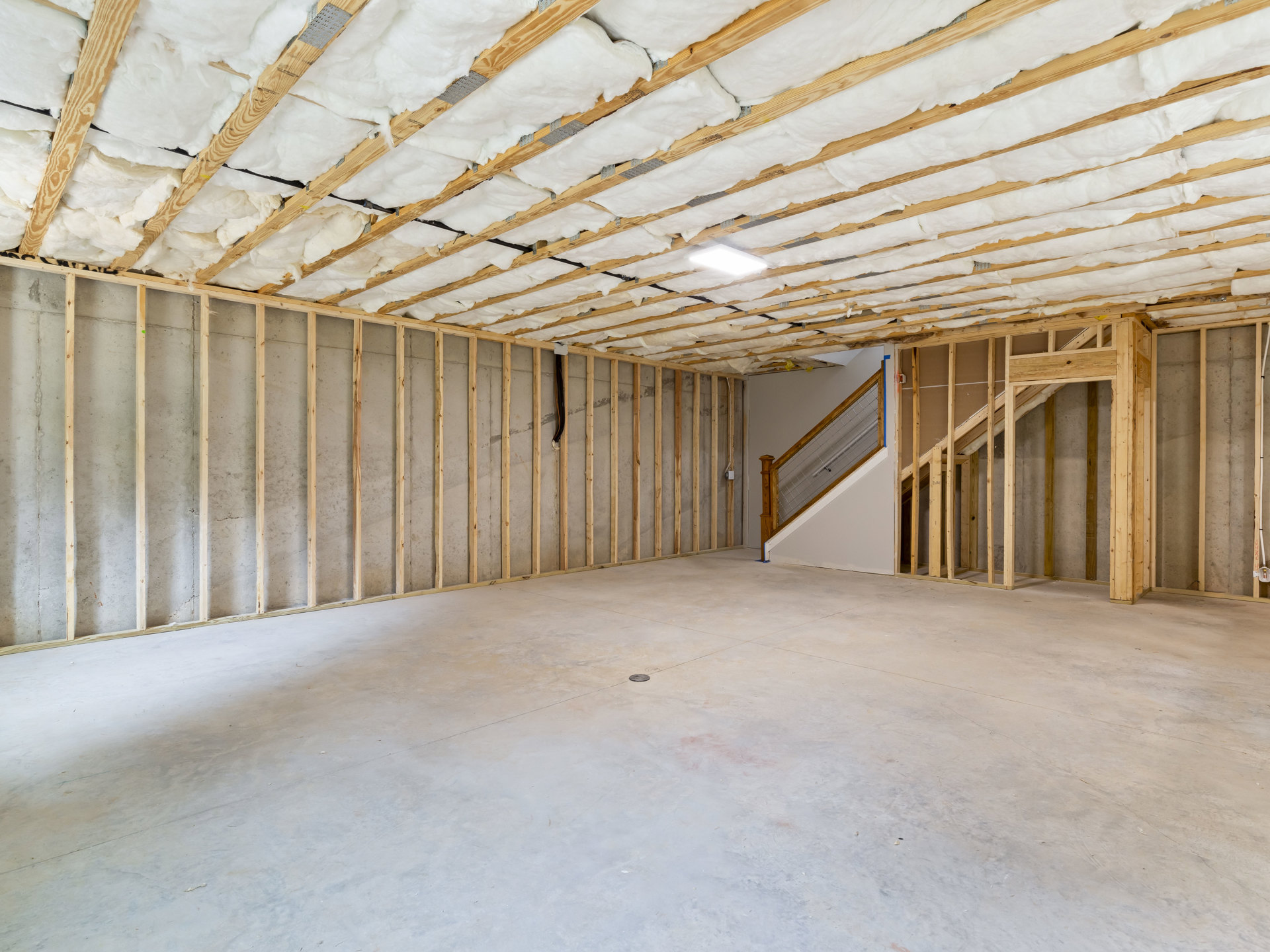 White ceiling with foam insulation, exposed wooden beams, metal staircase railing, concrete floor with drain, and white walls in a modern interior room.