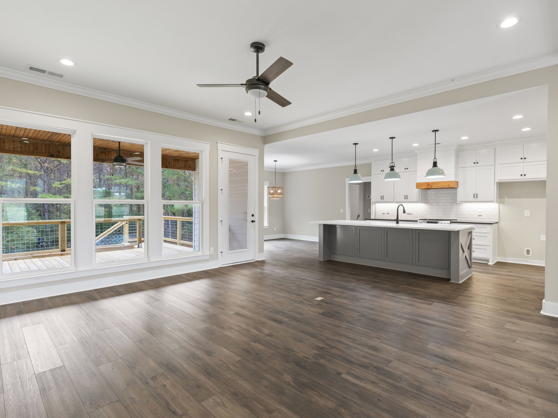 Spacious kitchen and dining area with wood flooring, white kitchen island topped with black pendant lamp, cabinetry along the wall, large windows overlooking outdoor grill.