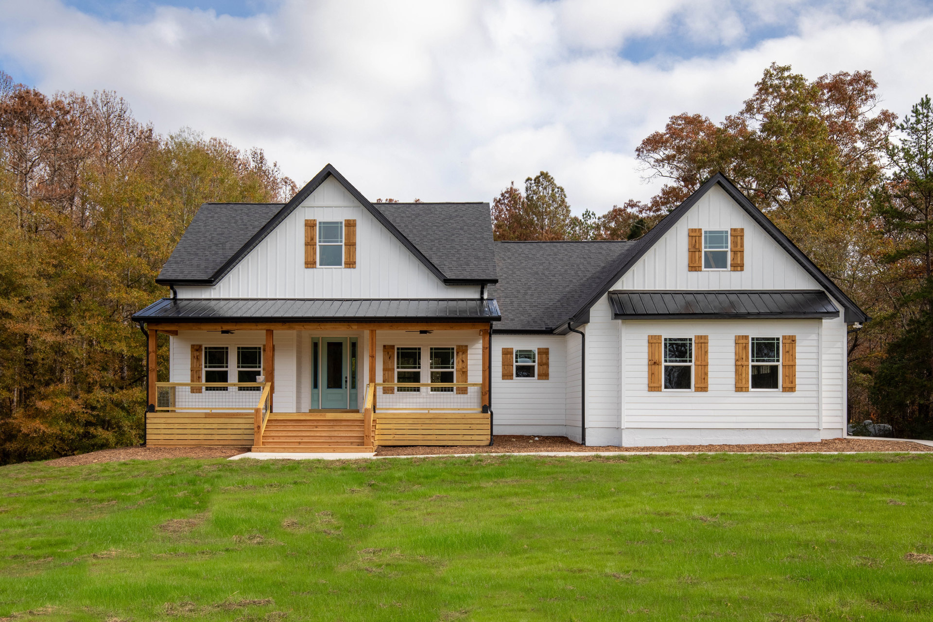 Two-story home with white siding, green front door with glass panels, covered porch, white-framed windows, manicured lawn, wooden fence, and mature trees in the background