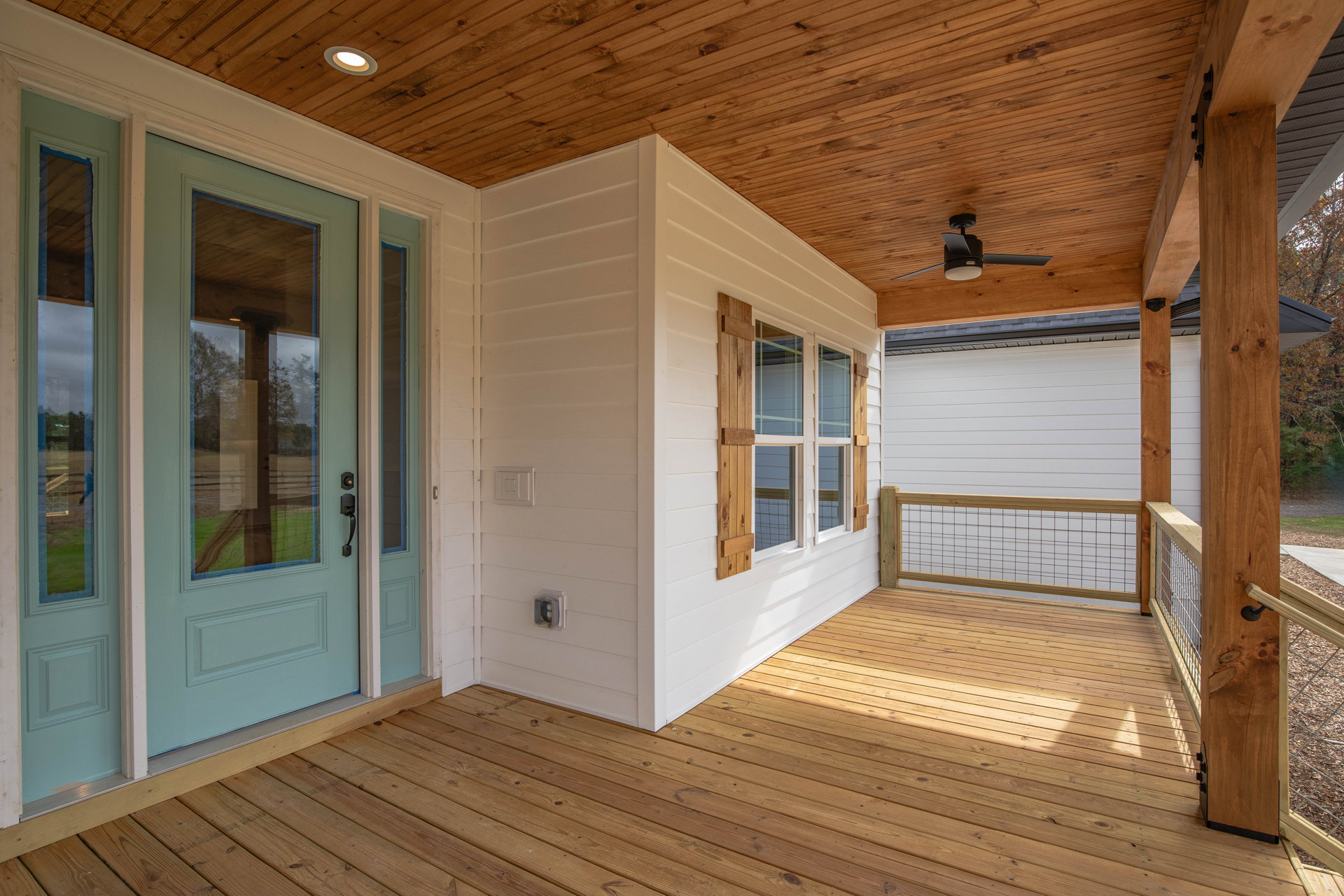 Wood deck porch with blue door, white wall, ceiling fan with light, wooden fence with wire mesh, window featuring wood shutters, and building corner visible