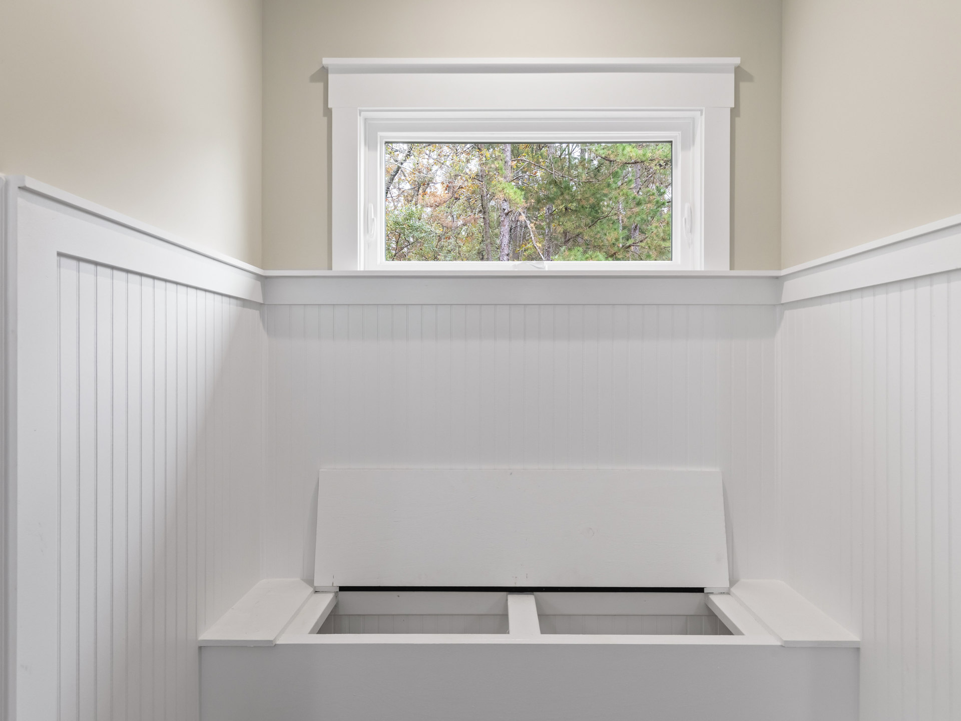 Wooden bench beneath large window with tree views, white walls with black striped accent, built-in white shelf, decorative molding.