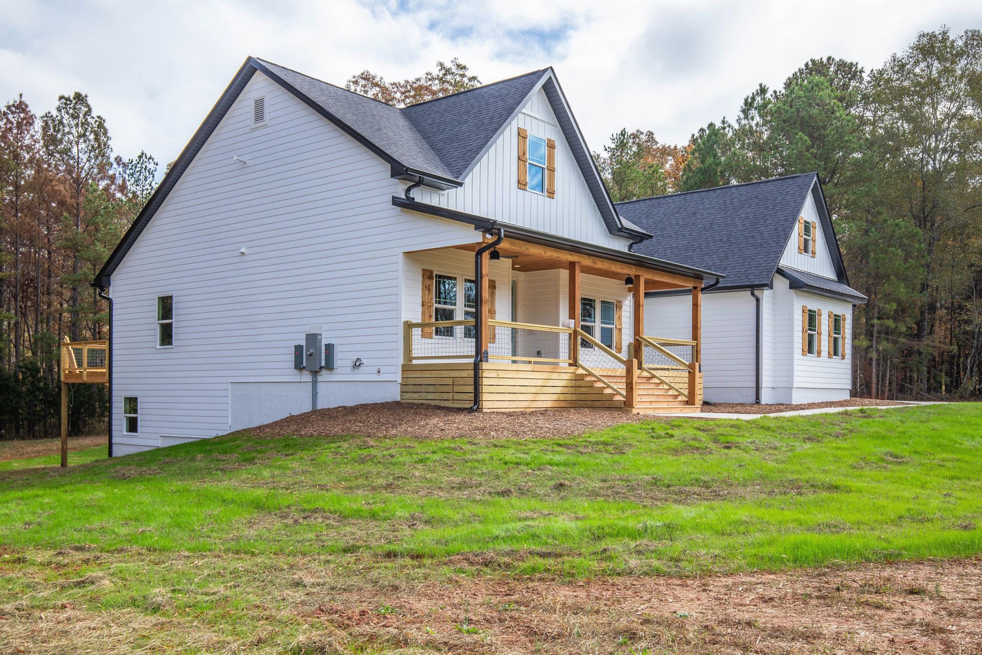 Two-story cottage with wood siding, covered front porch, wire railing, manicured lawn, large windows reflecting trees, rope detail tied to porch post, partly cloudy sky overhead