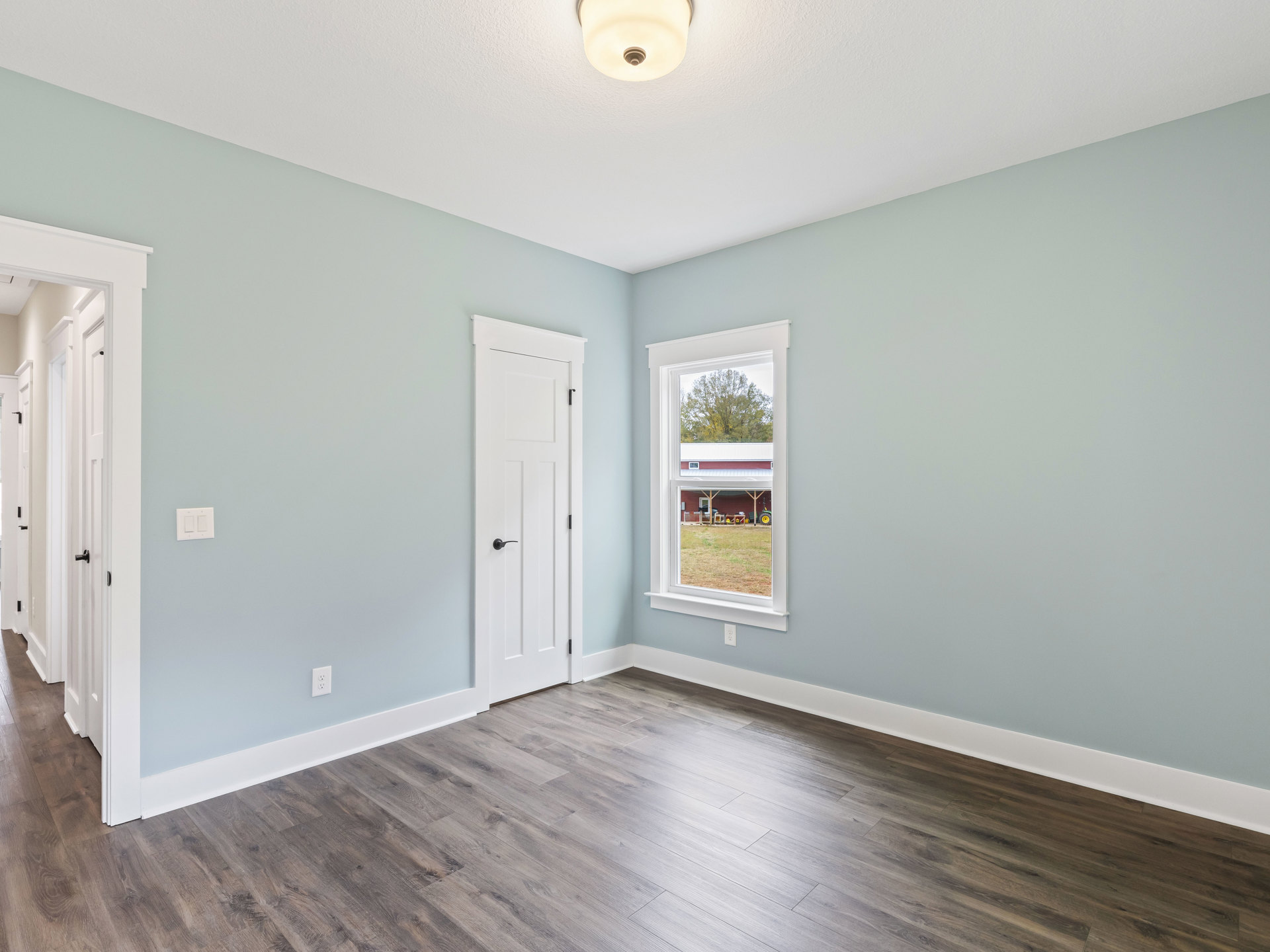 White door with black handle, wood flooring, round ceiling light fixture, window overlooking farm and tractor, plaster walls