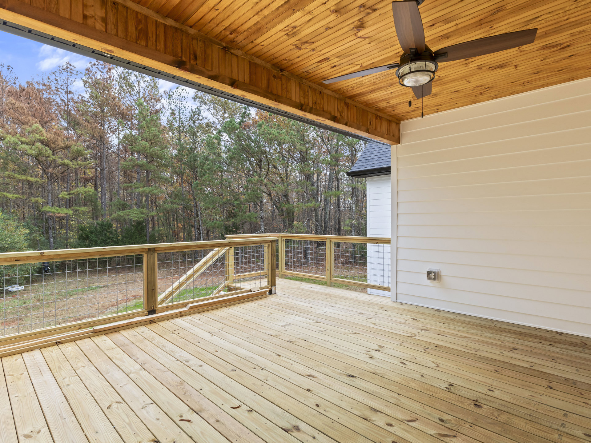 Wooden deck with metal railings, white wall, wood plank ceiling, ceiling fan, and trees visible in the background