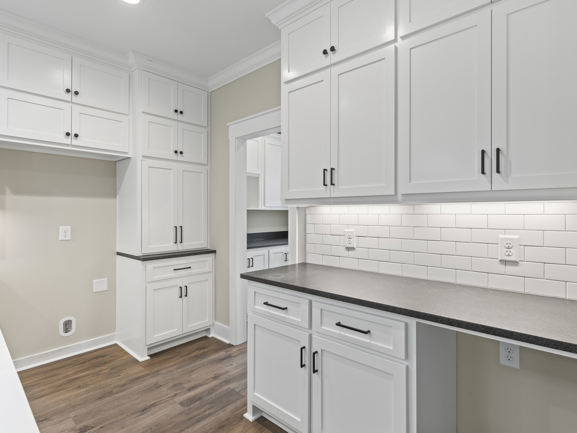Kitchen with white cabinets, black countertops, wood flooring, black cabinet handles, white door with black handle, and visible electrical outlet