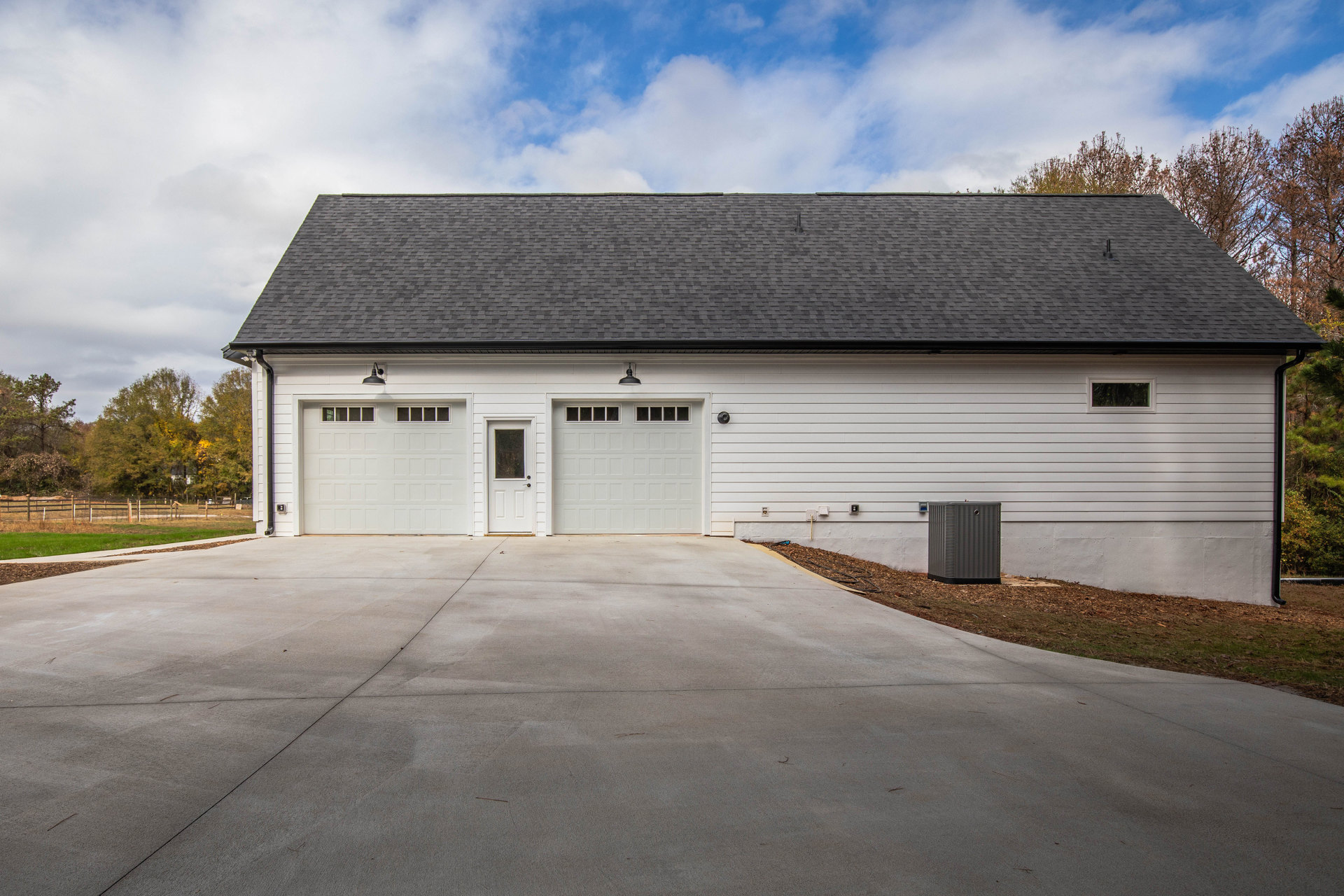 White garage with black roof, paneled garage door featuring rectangular windows, surrounded by trees and cloudy sky