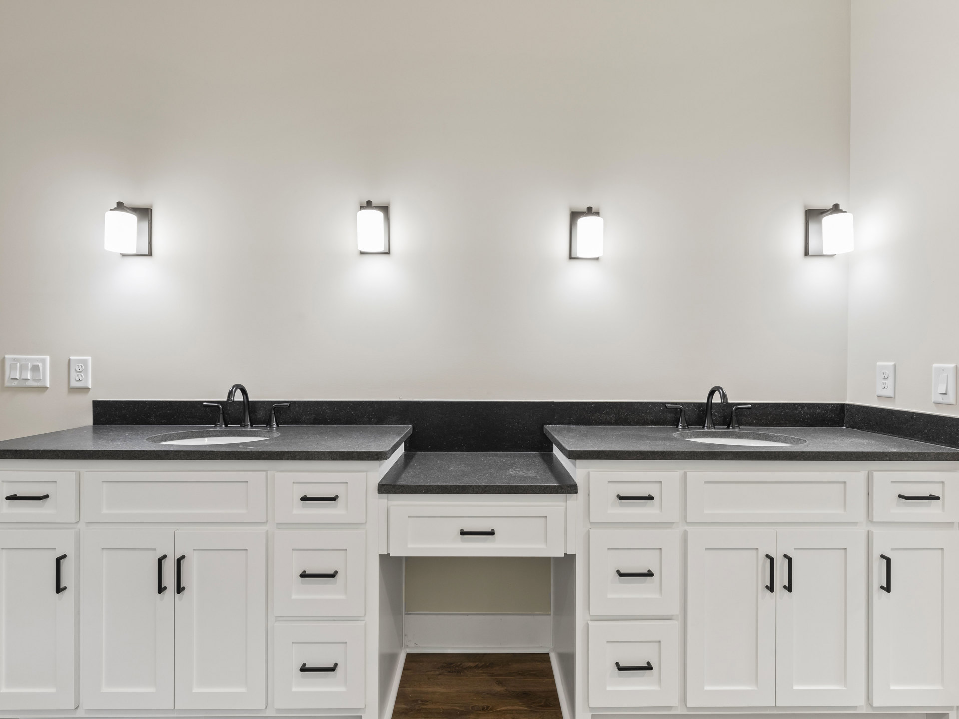 White bathroom cabinets with matte black countertops, chrome faucet, wall-mounted light fixture, wood drawer fronts, and a white light switch.