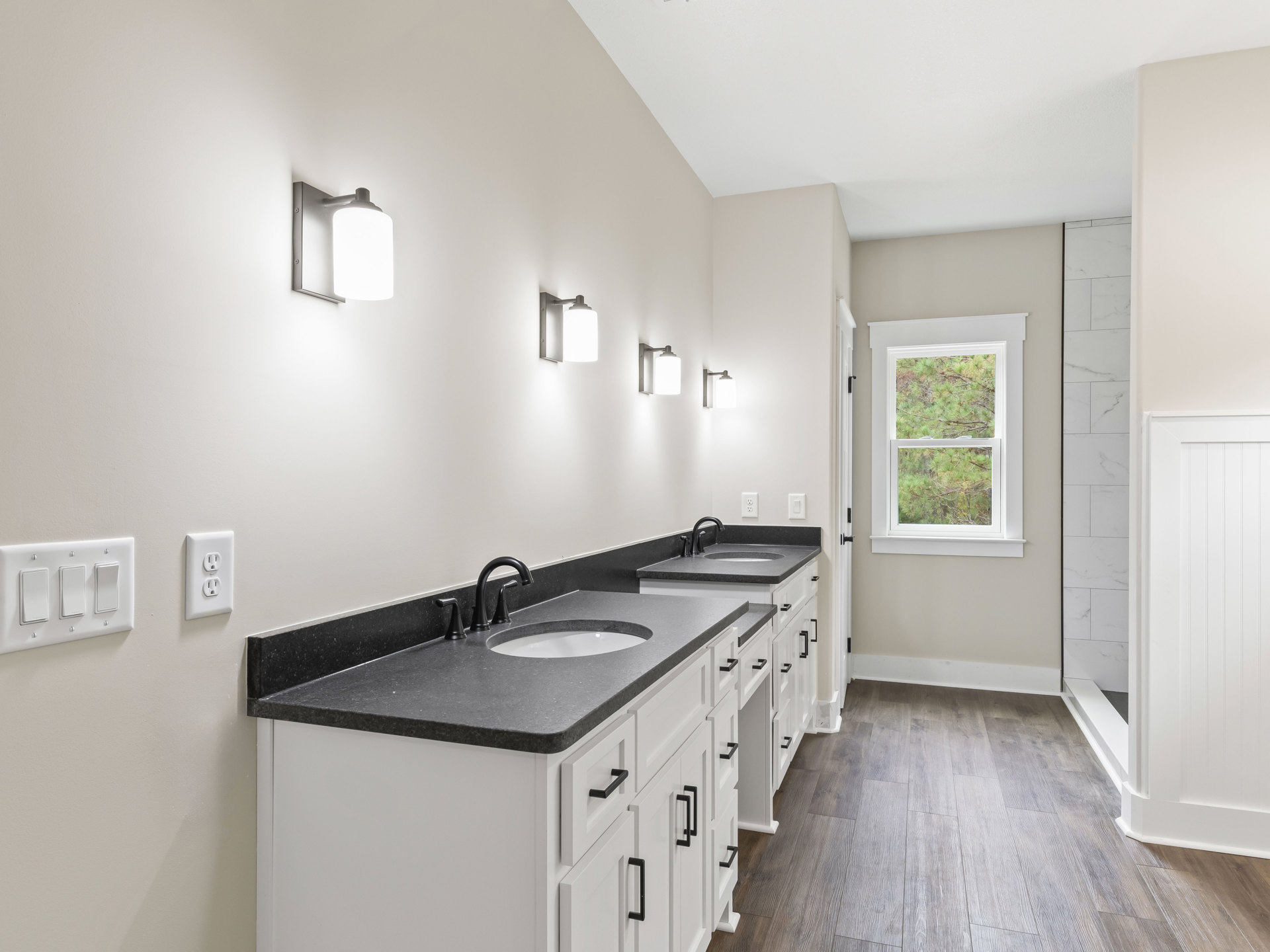Bathroom featuring white cabinetry, black countertops, rectangular window overlooking trees, wall-mounted light fixture, white electrical outlet and light switch, close-up of