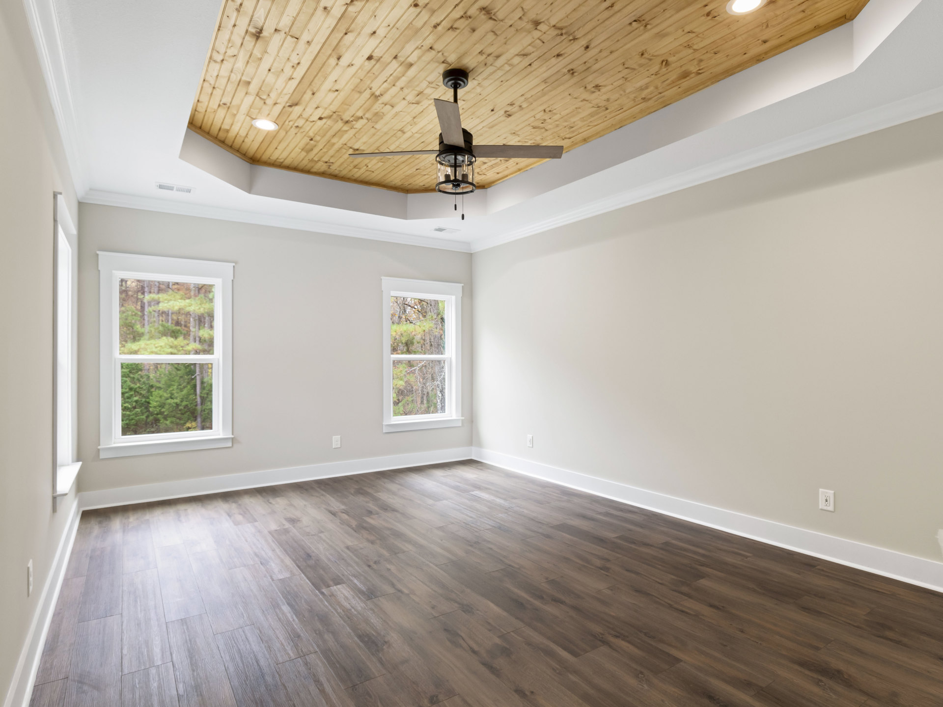 Wood-floored room with wood ceiling, ceiling fan with light fixture, large windows framing leafy trees outside, plaster walls, and decorative molding