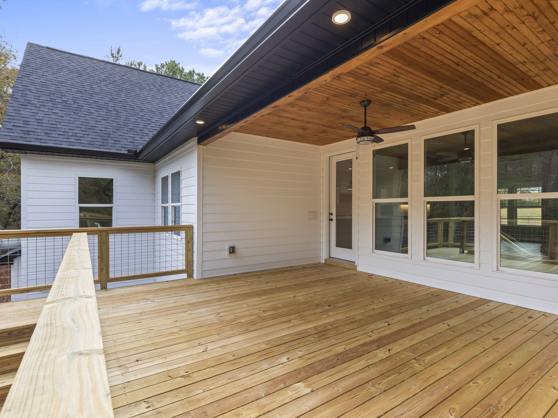 Covered deck with wood plank flooring, wood railing, white exterior wall, glass door, and two ceiling fans mounted on a wood-paneled ceiling