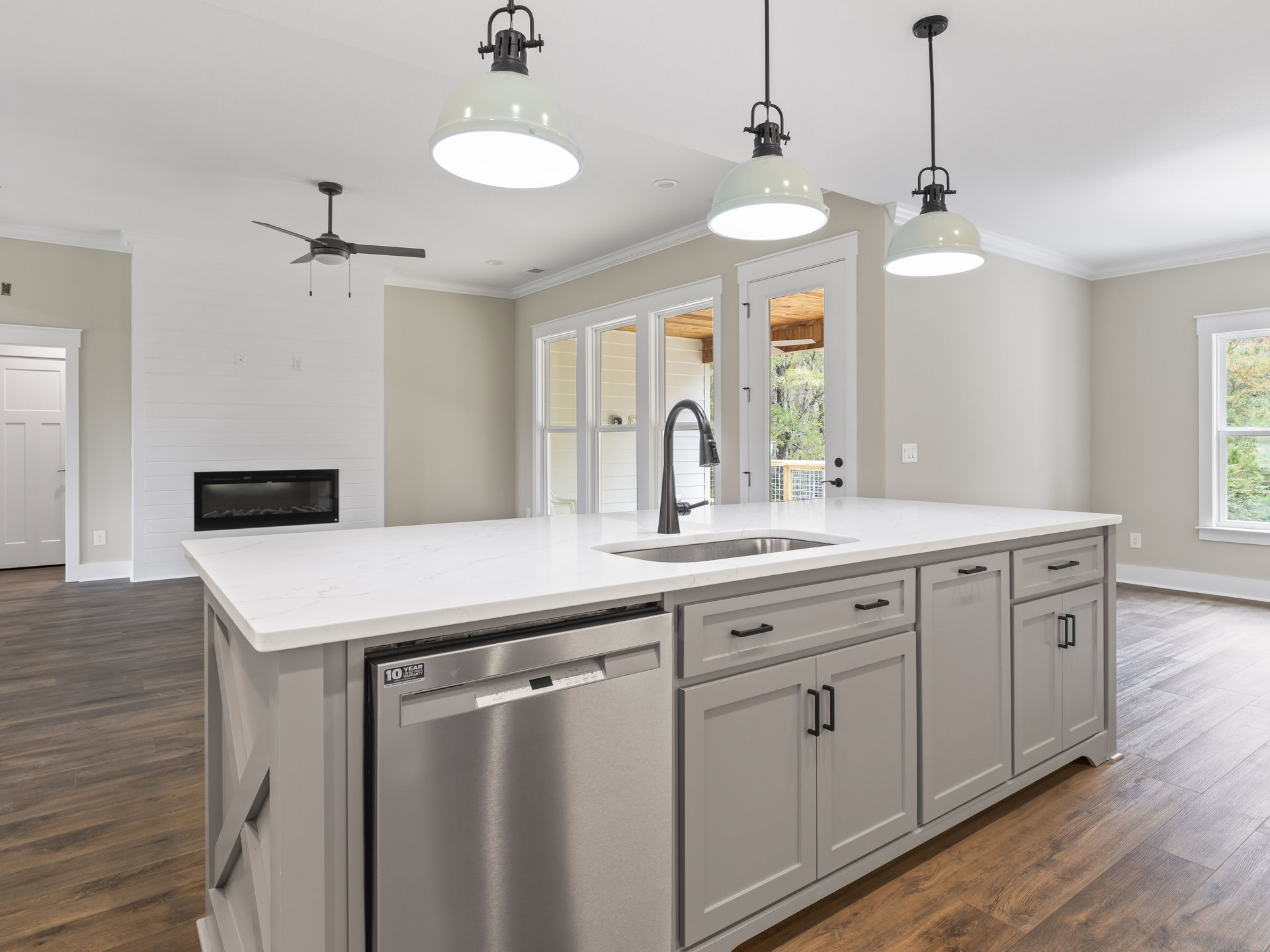 White kitchen cabinets, stainless steel dishwasher, black and white ceiling light fixture, window with view of trees, light countertops, wood flooring