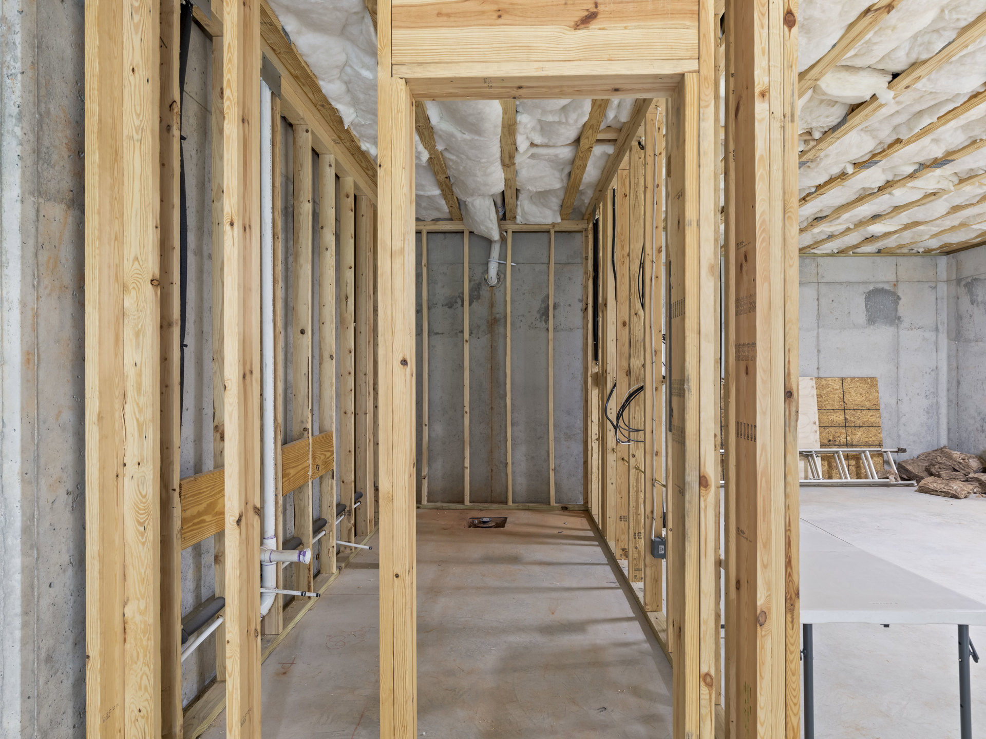 Unfinished room with exposed wood framing, white foam insulation on ceiling beams, grey wall with paint spot, and ladder leaning against wall