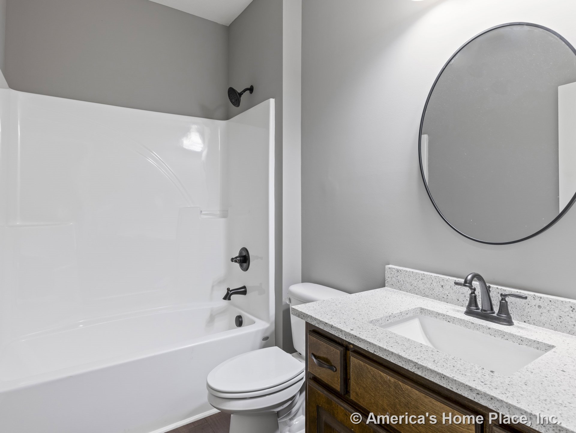 White acrylic tub-shower combo with granite vanity countertop, dark wood cabinetry, matte black faucet, round wall mirror, gray painted walls, and built-in lighting.