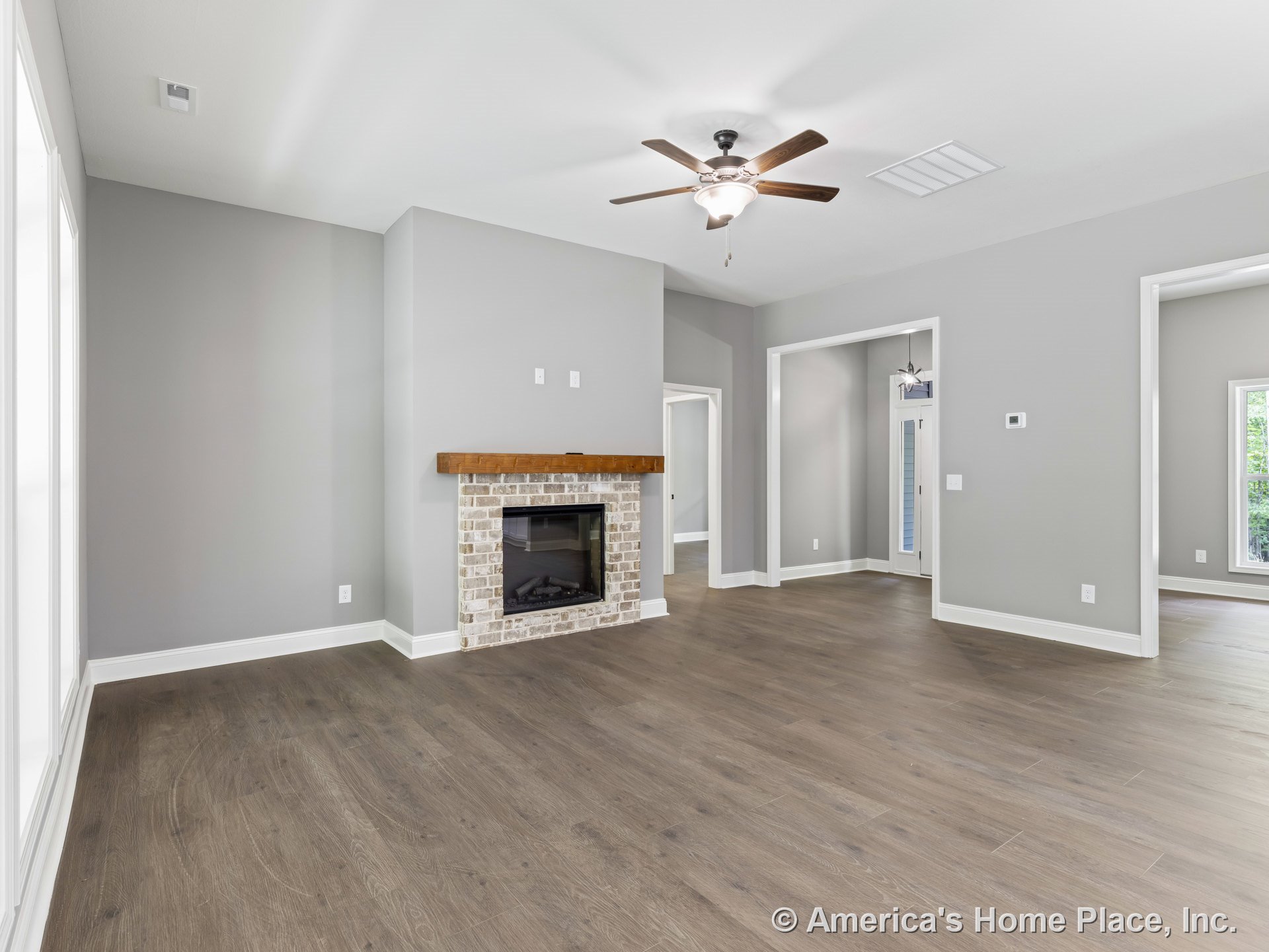 Brick fireplace with wooden mantel, wide plank wood flooring, gray painted walls, white trim and baseboards, ceiling fan with light fixture, large windows, and adjacent doorways.