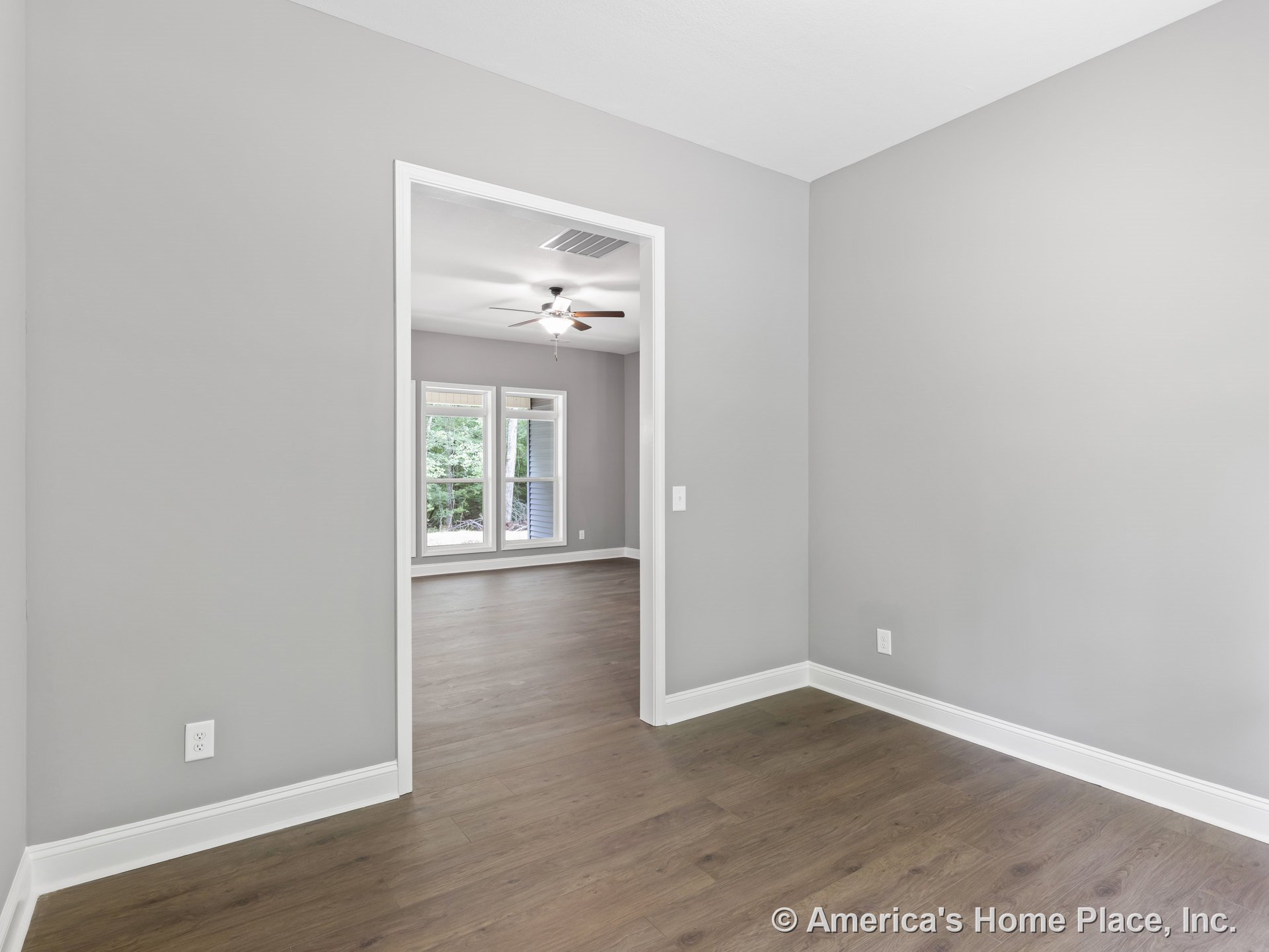 Open doorway connecting living room and bedroom with wood-look flooring, gray painted walls, white baseboards and trim, large windows, ceiling fan with light fixture, and abundant