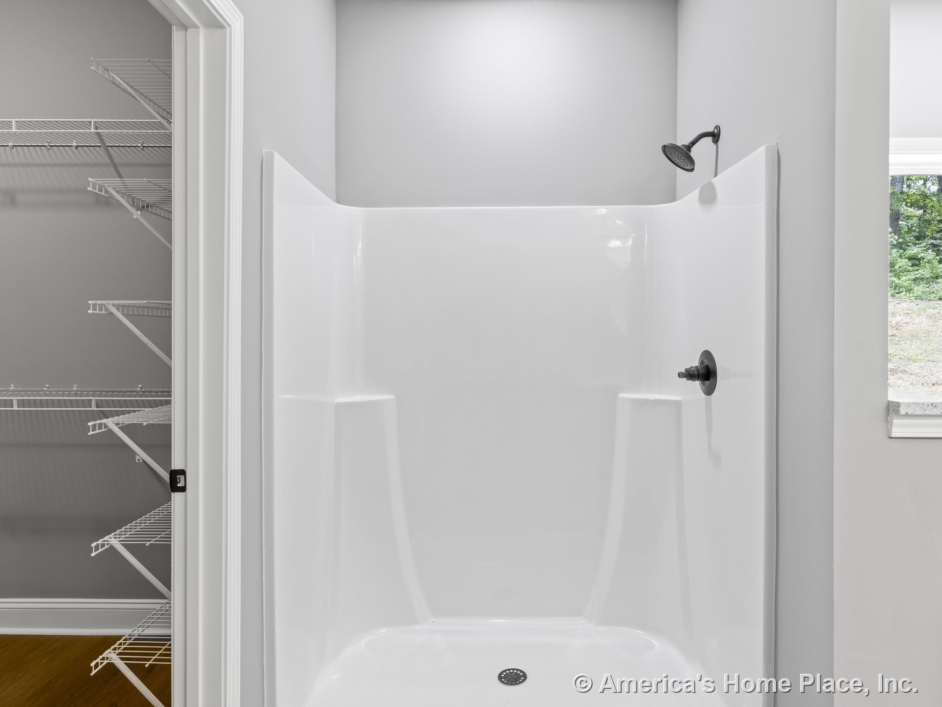 White fiberglass shower enclosure with integrated shelving and matte black showerhead and handle, adjacent wire shelving closet, white trim and door frame, window providing natural