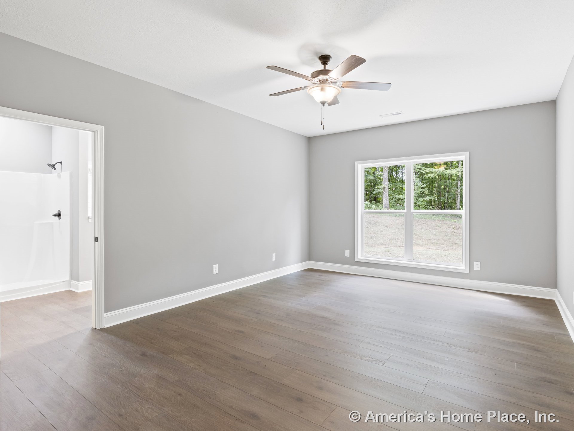 Bedroom with wide plank wood flooring, gray painted walls, white trim and baseboards, double window with white casing, ceiling fan and light fixture, and attached bathroom